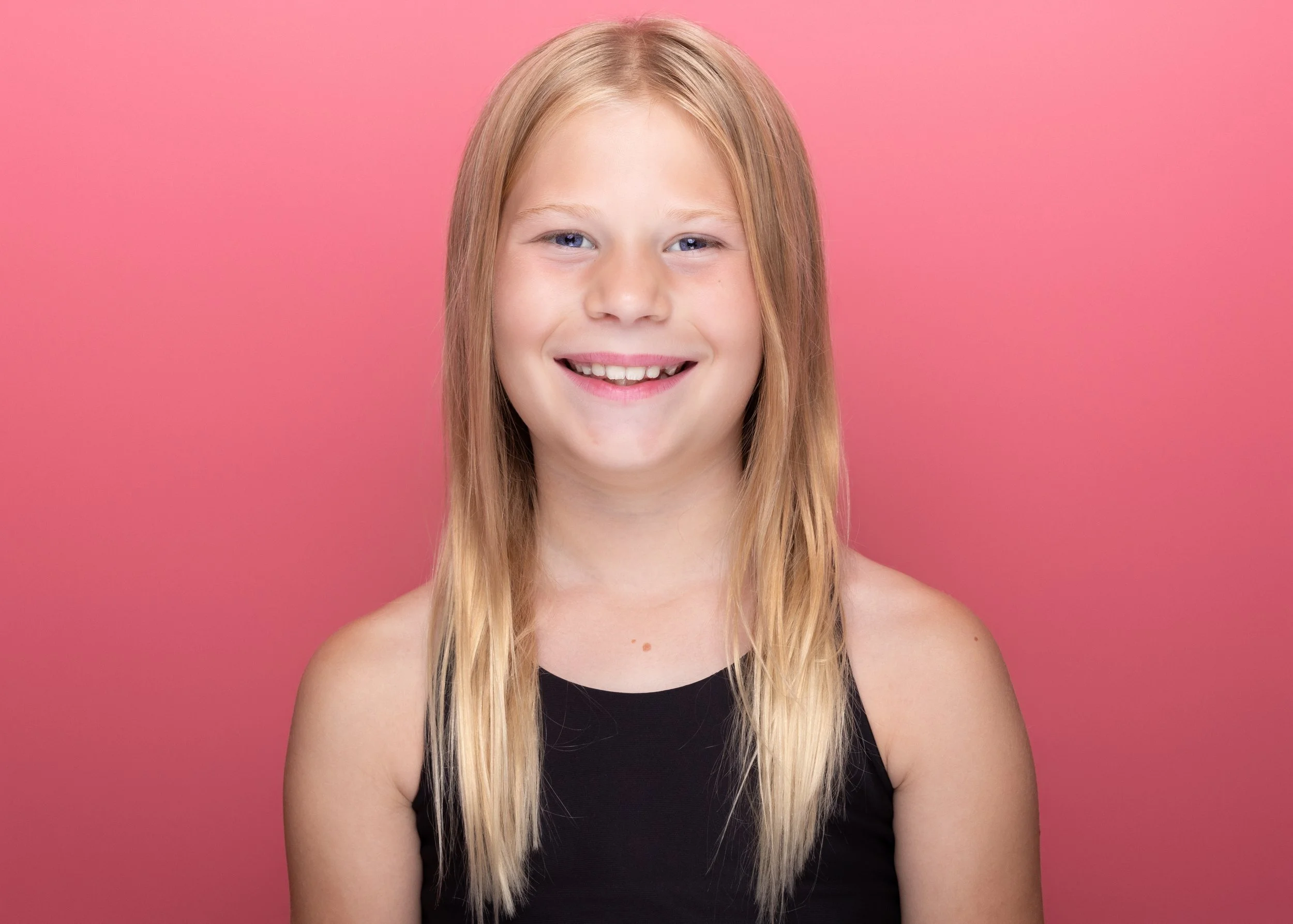 A young girl with long blonde hair, blue eyes, and light skin, smiling, wearing a black top, against a pink background.