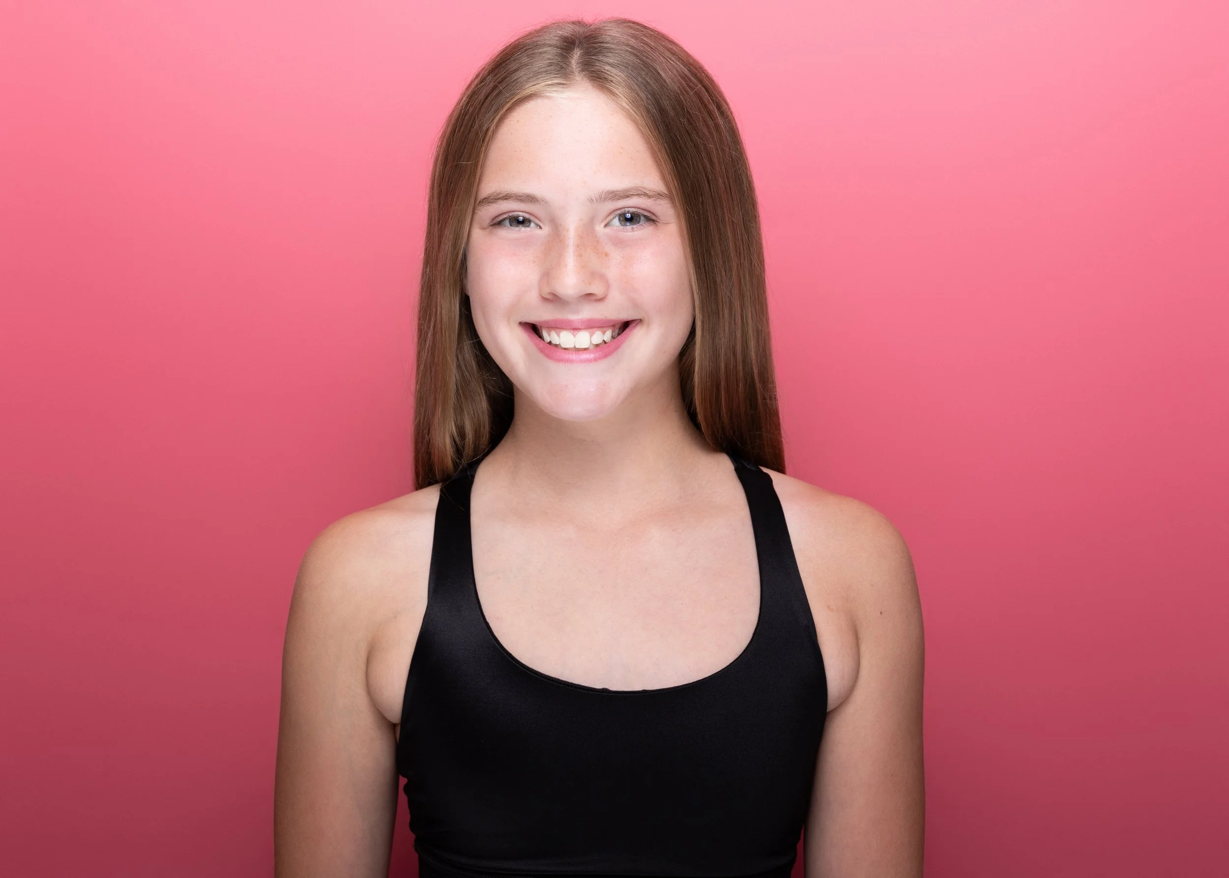 A young woman with long red hair, smiling, wearing a black tank top, standing against a pink background.