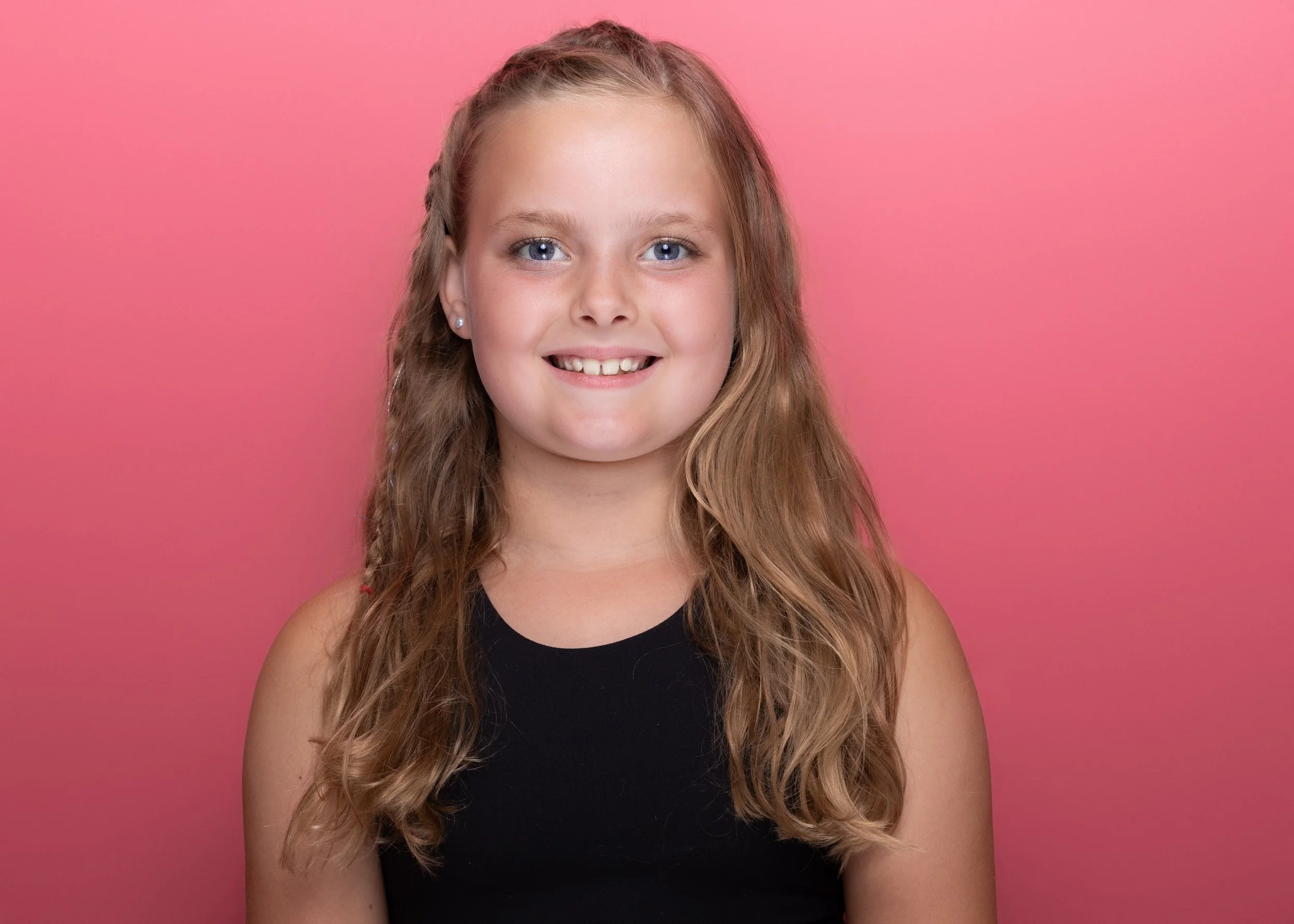 A young girl with long, wavy light brown hair and blue eyes, smiling, wearing a black tank top, against a pink background.