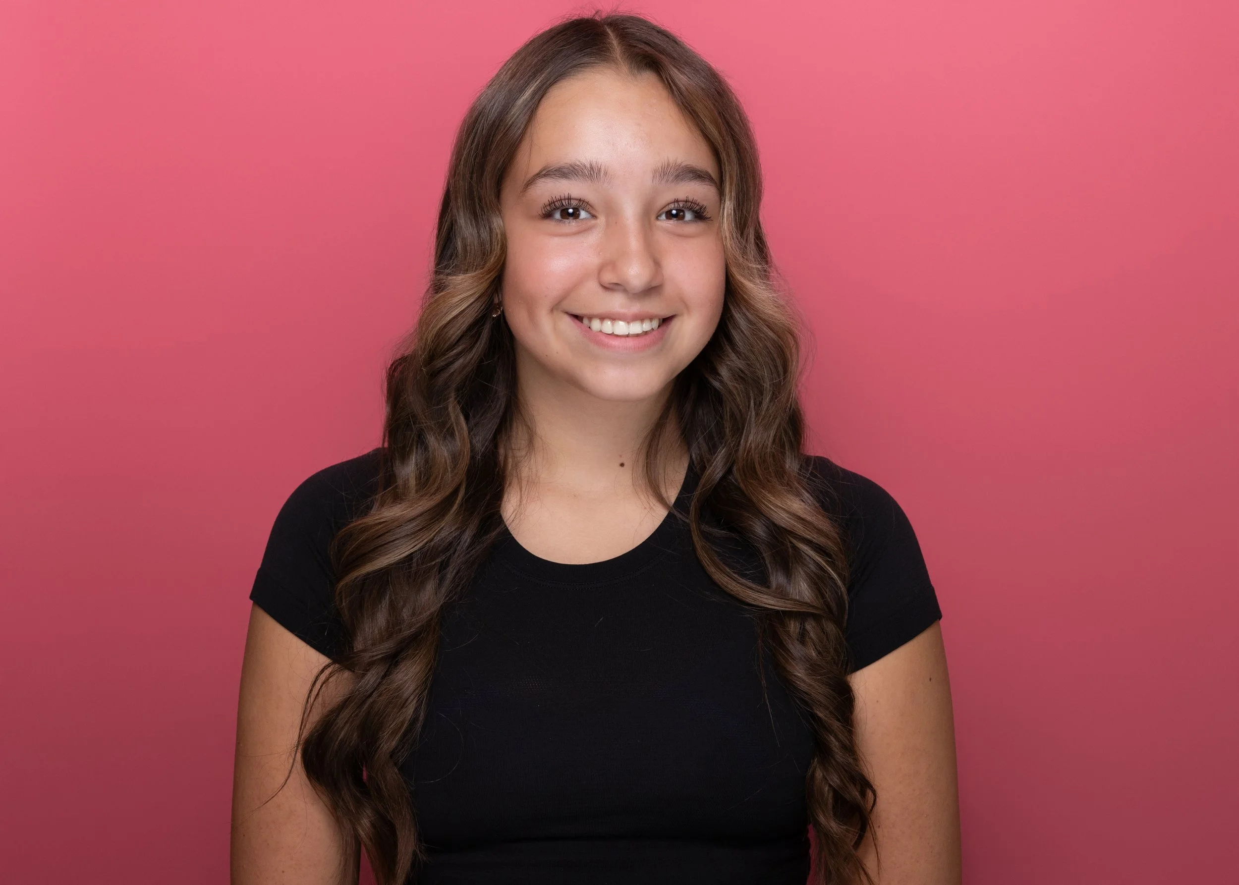 A young woman with long brown curly hair, wearing a black t-shirt, smiling against a pink background.
