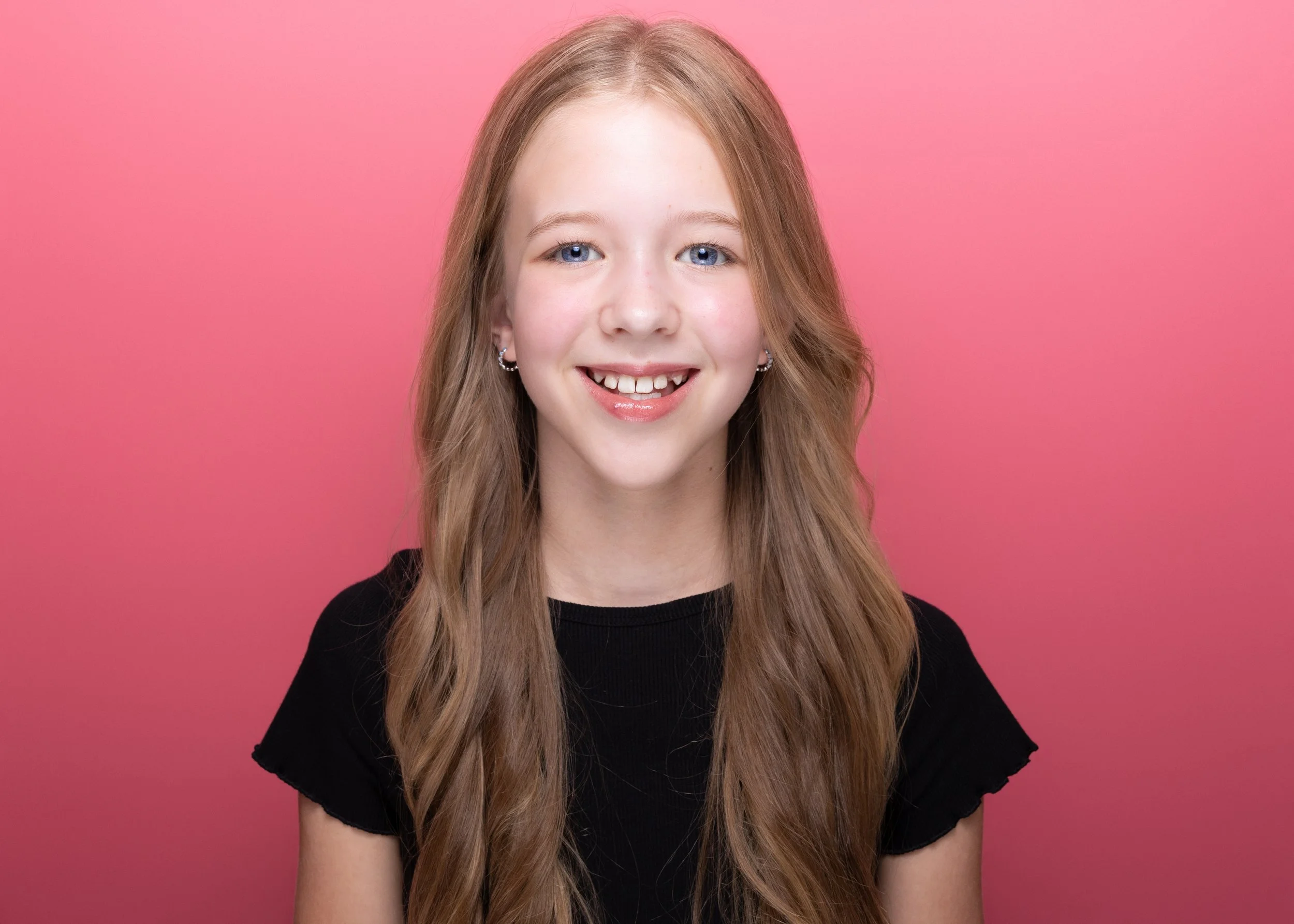 A young girl with long, wavy blonde hair and blue eyes, smiling, wearing a black shirt, against a pink background.