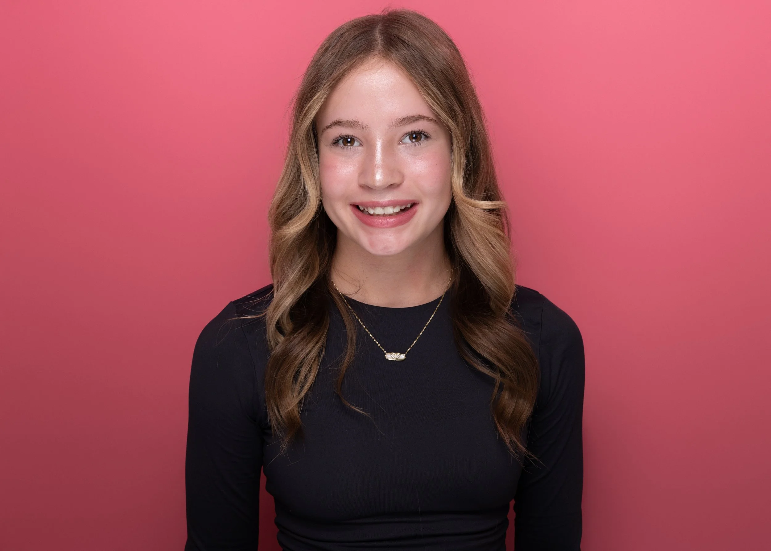 A young woman with long wavy hair smiling, wearing a black top and a necklace, standing against a pink background.