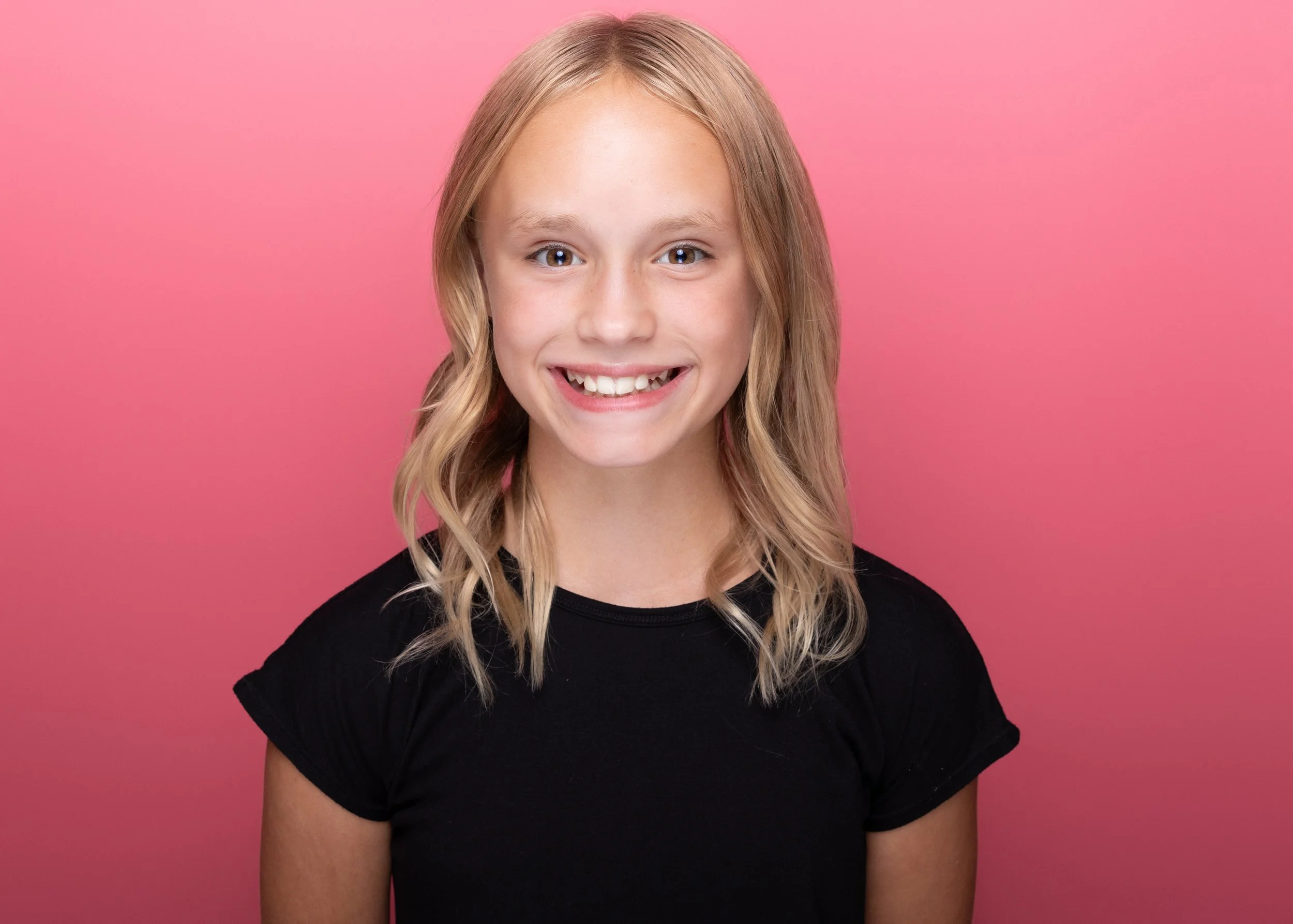 A young blonde girl with wavy hair, smiling, wearing a black T-shirt, against a pink background.