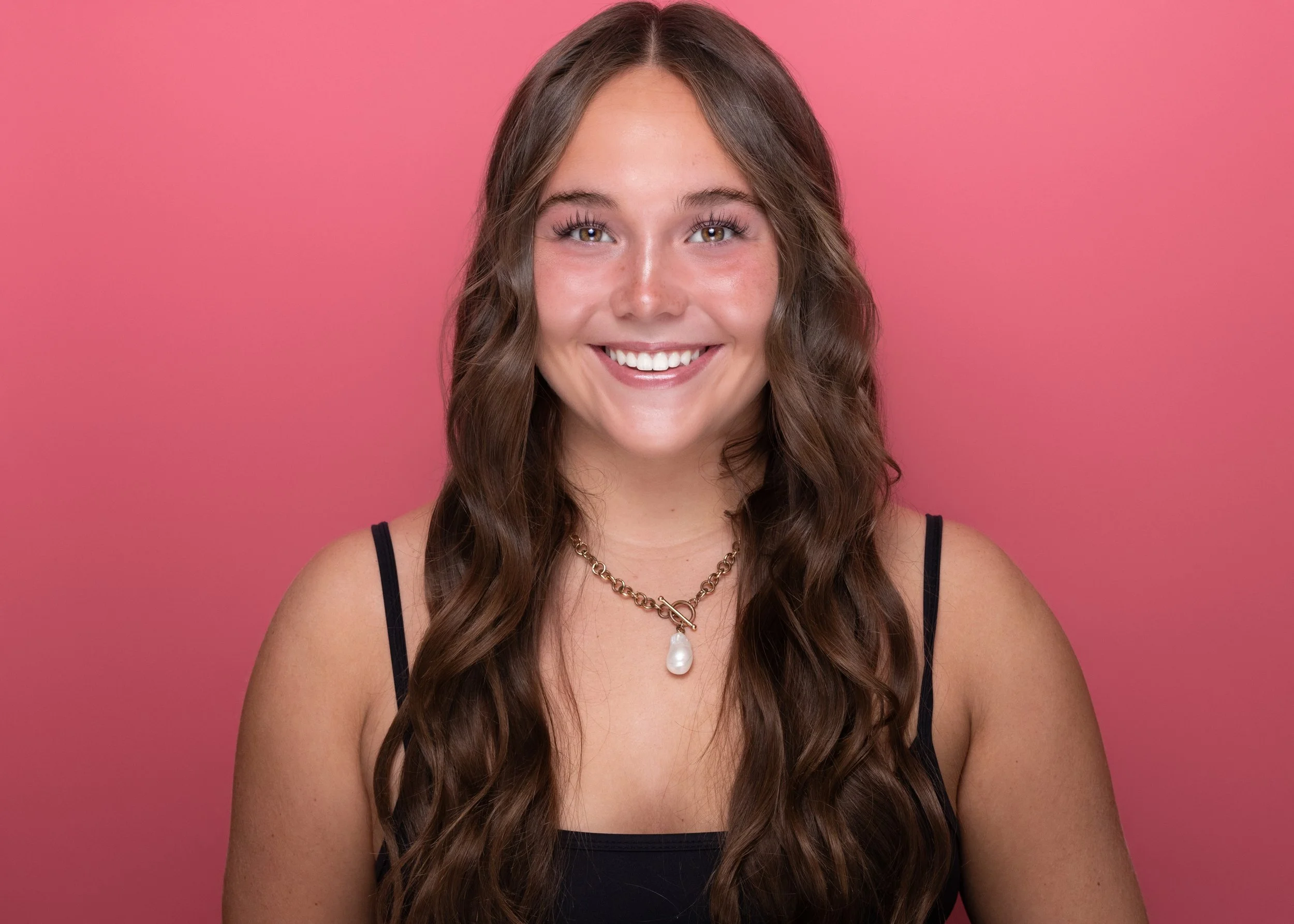 A young woman with long, wavy brown hair smiling against a pink background. She is wearing a black tank top, a gold chain necklace, and a large pearl pendant.