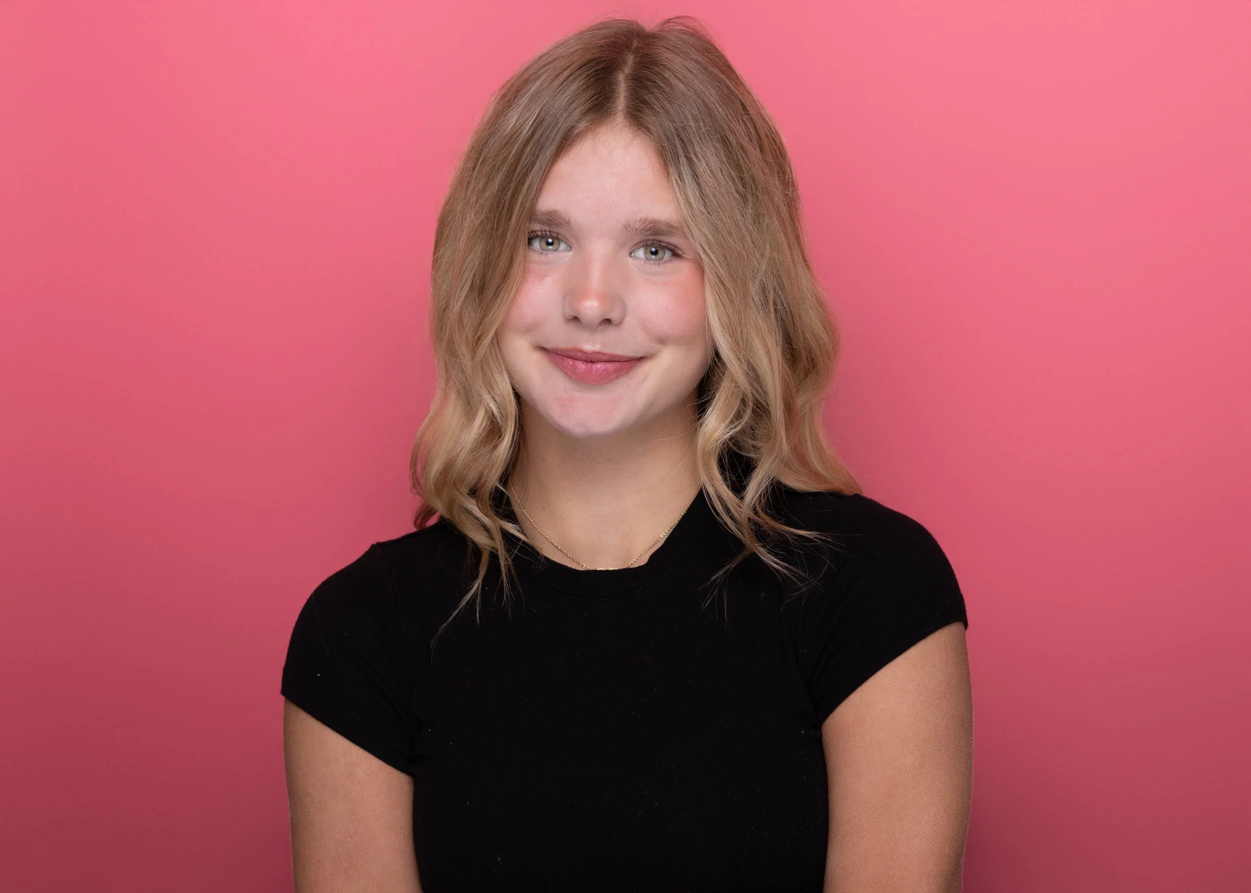 A young woman with blonde, wavy hair and light blue eyes smiling, wearing a black shirt and a gold chain necklace, against a solid pink background.