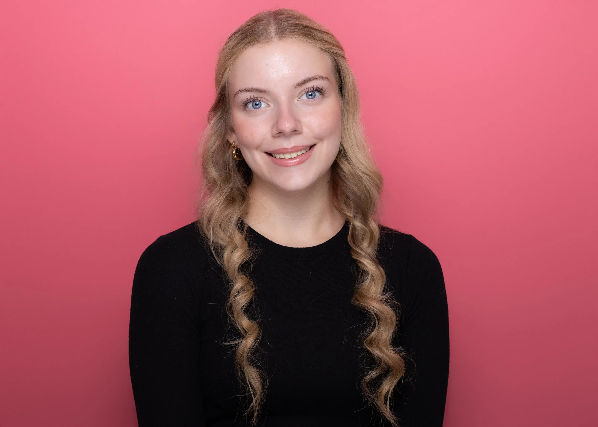 A young woman with blonde hair, blue eyes, and gold earrings, smiling while looking at the camera, against a pink background, wearing a black top.