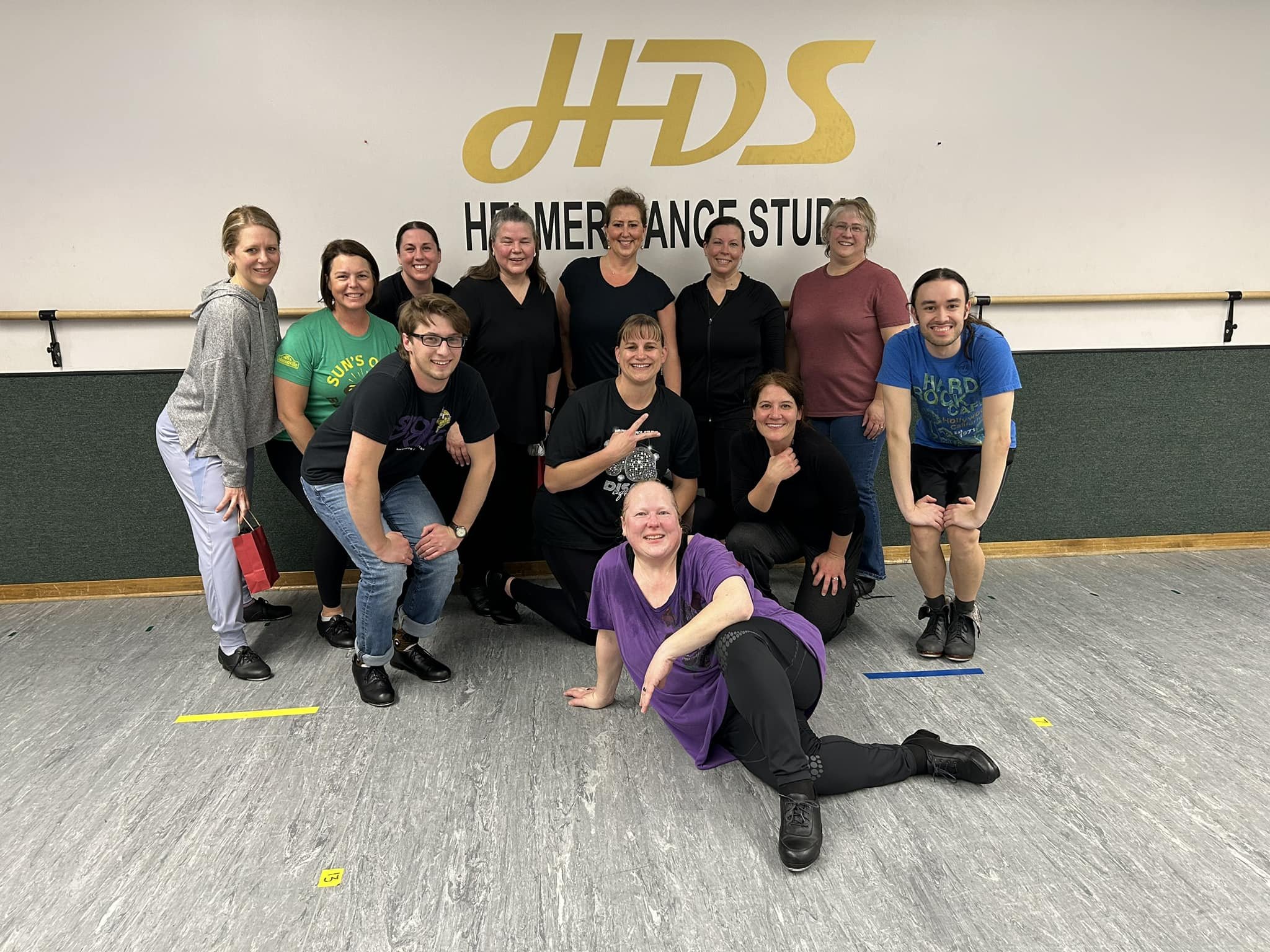 Group of people standing in a dance studio in front of a wall with the logo 'HHS' and the words 'Homer Performance Studio'. Some individuals are kneeling in front, some are standing, and they are smiling at the camera.