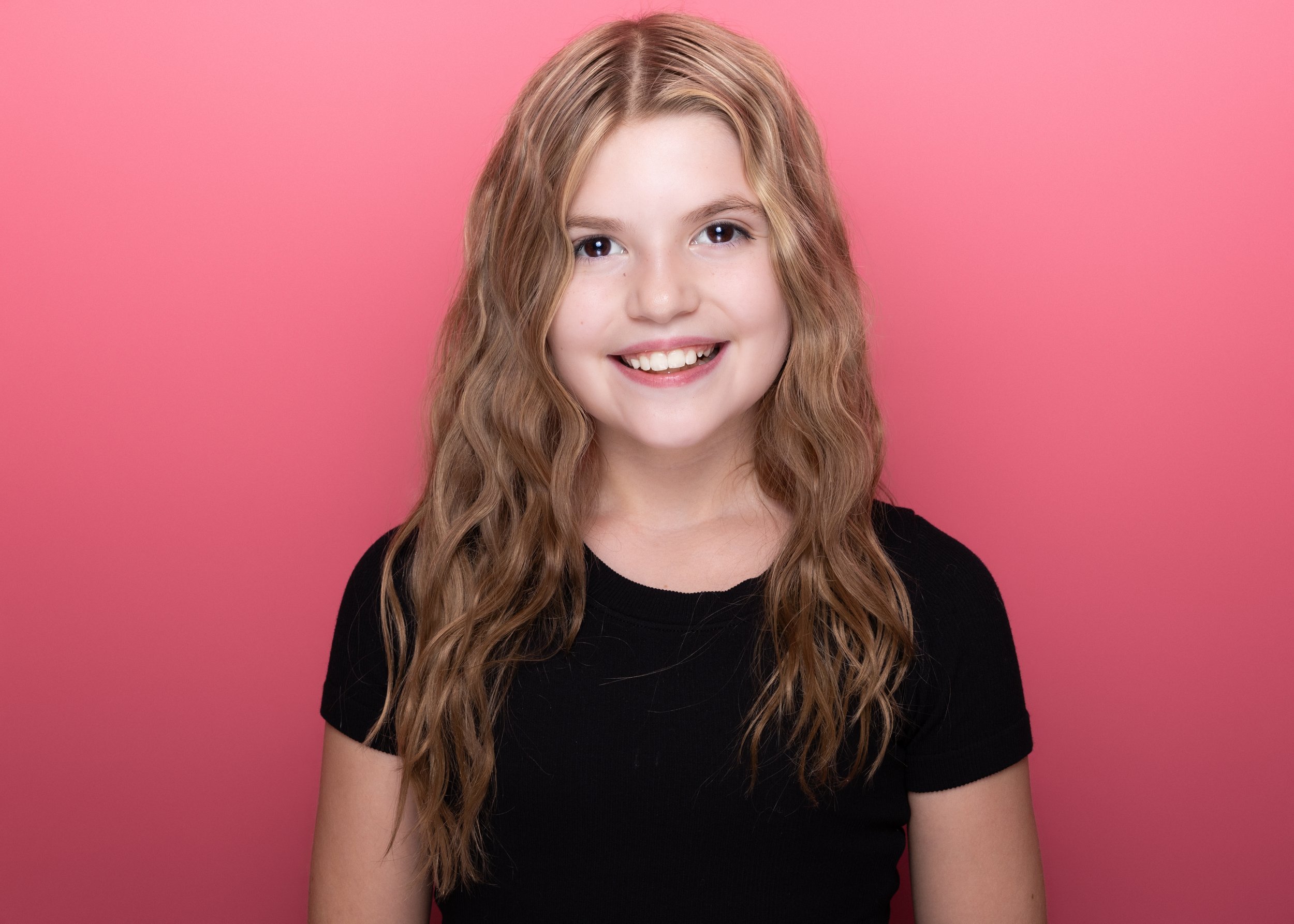 A young girl with long wavy blond hair, smiling showing her teeth, wearing a black t-shirt, standing in front of a solid pink background.