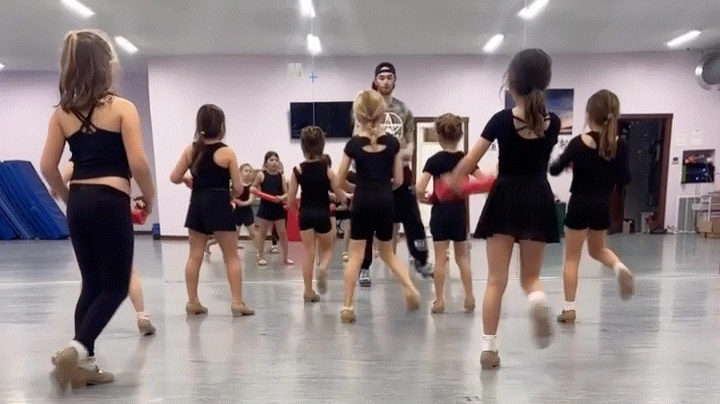 Young girl in black dance outfit holding hands with her dance instructor during rehearsal inside a dance studio. Other young dancers practice in the background.