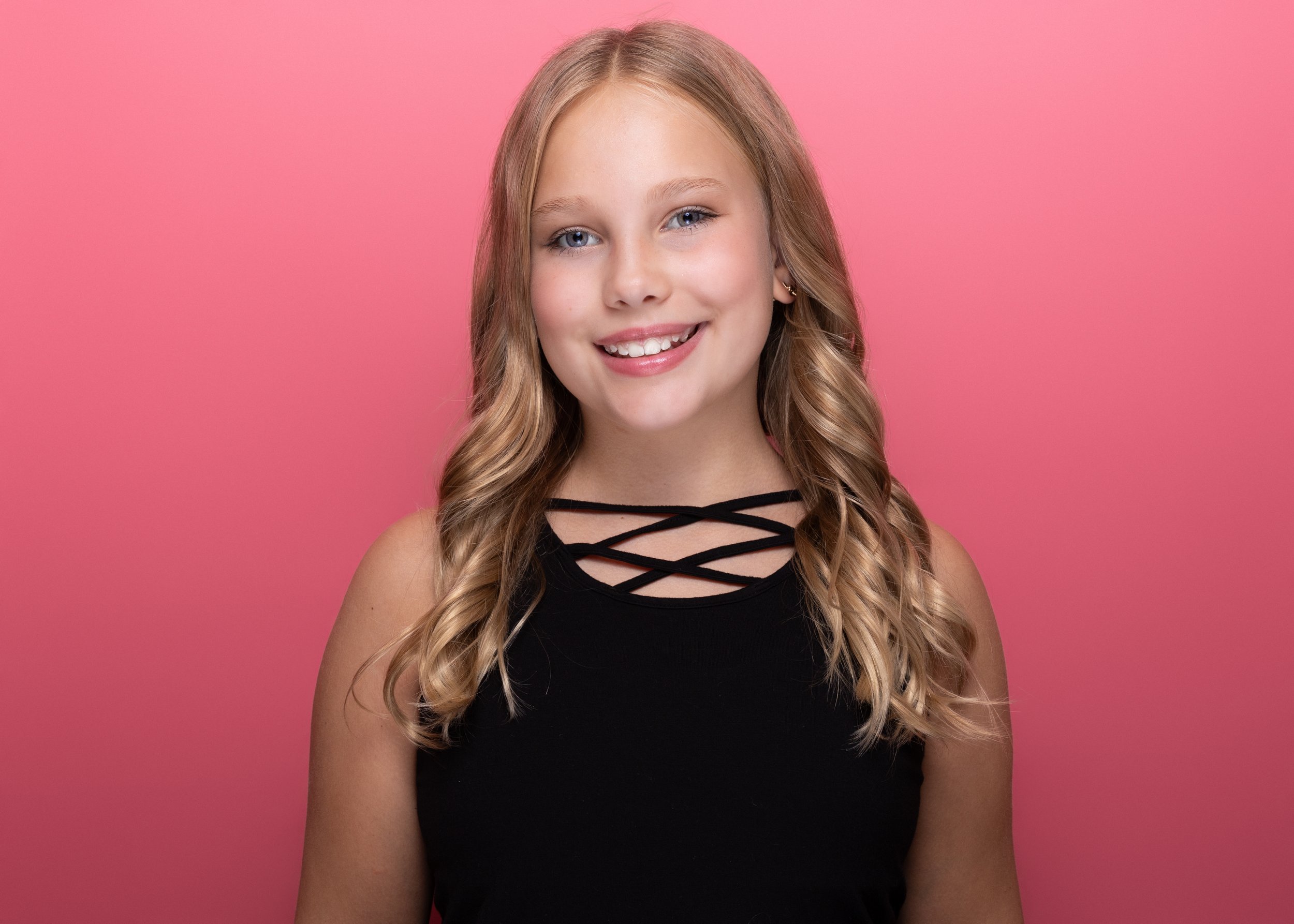 A young girl with long wavy blonde hair, wearing a black sleeveless top with crisscrossed straps near the neckline, smiling and showing her teeth, standing against a pink background.
