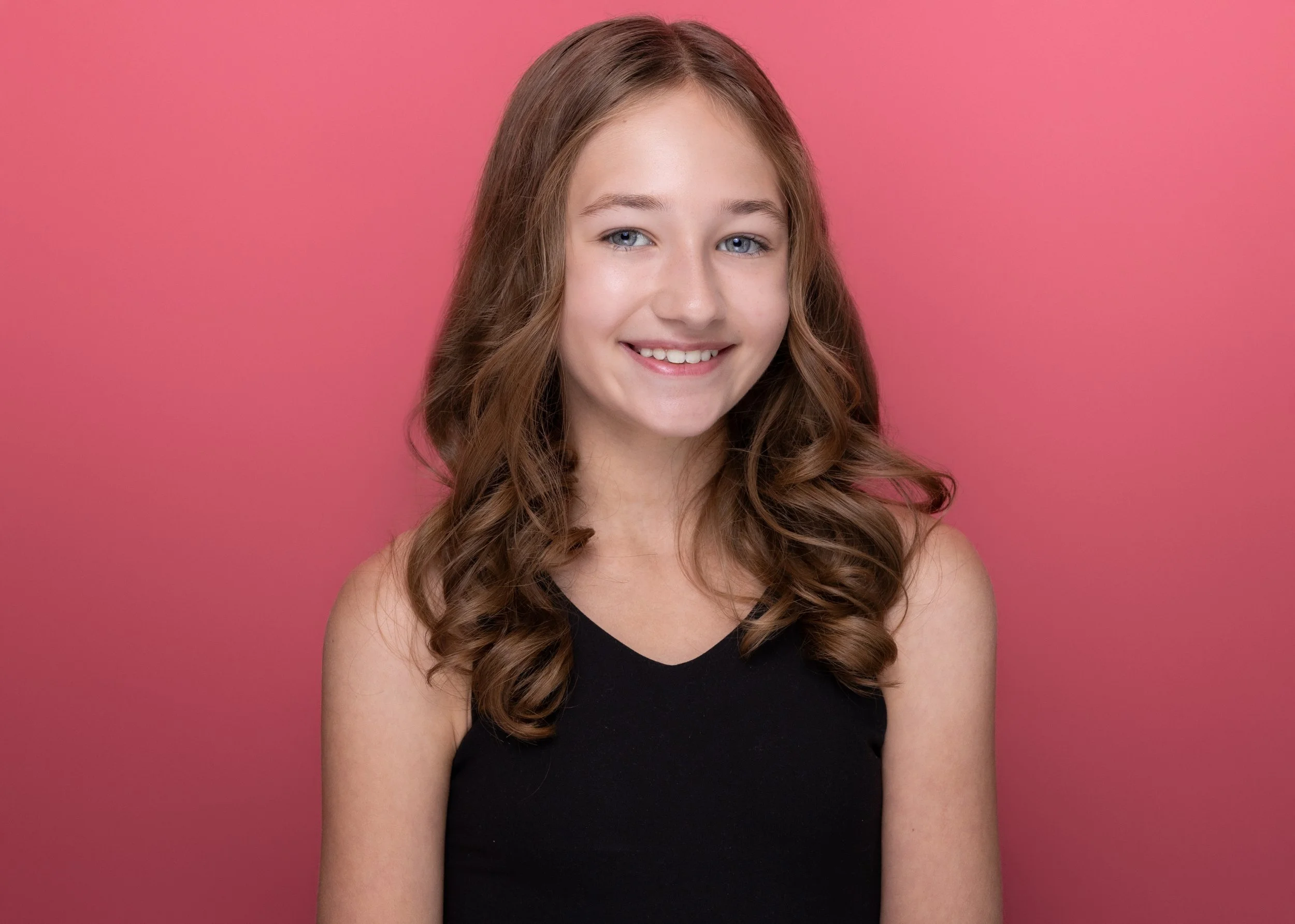 A young woman with long, curled brown hair and blue eyes smiling, wearing a black sleeveless top, against a pink background.