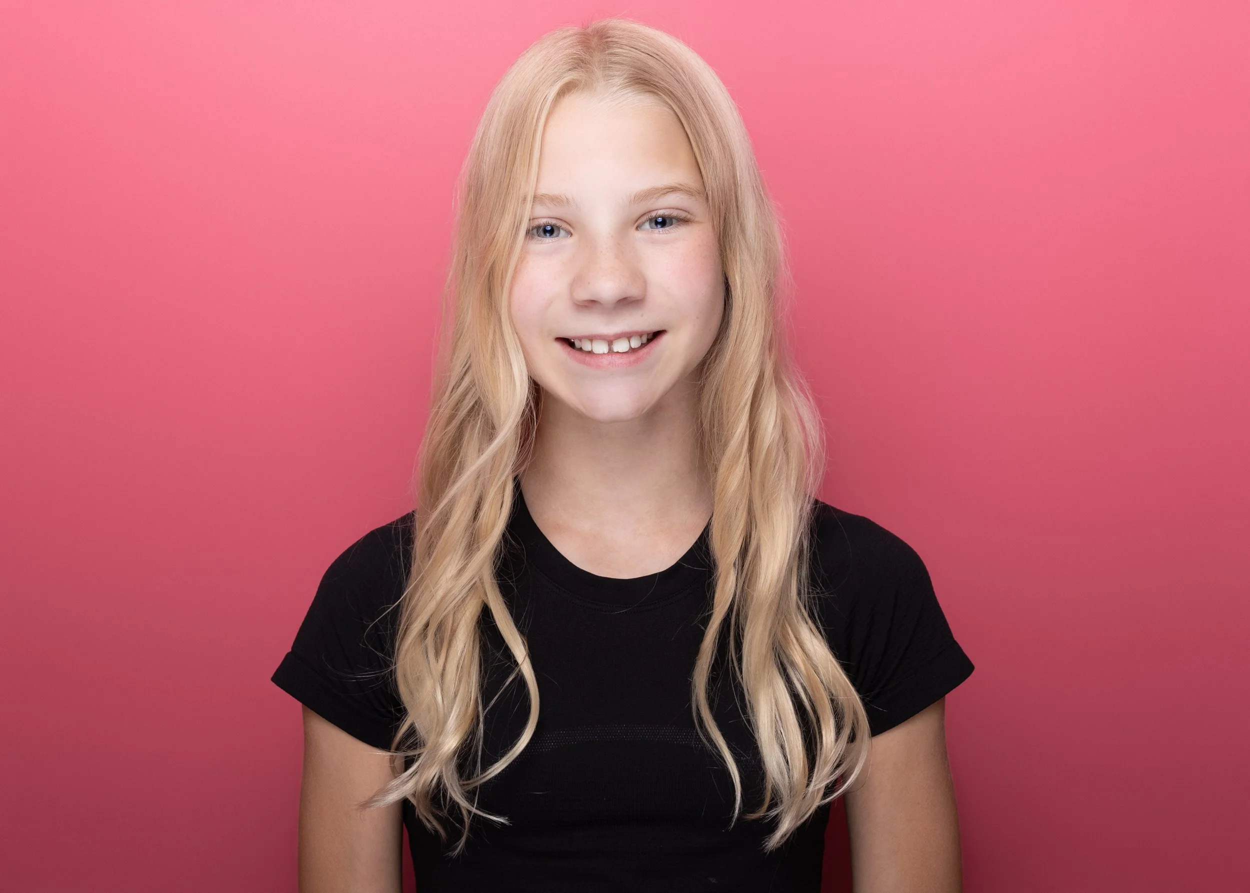 A young girl with long blond hair, blue eyes, and freckles, wearing a black shirt, smiling in front of a pink background.