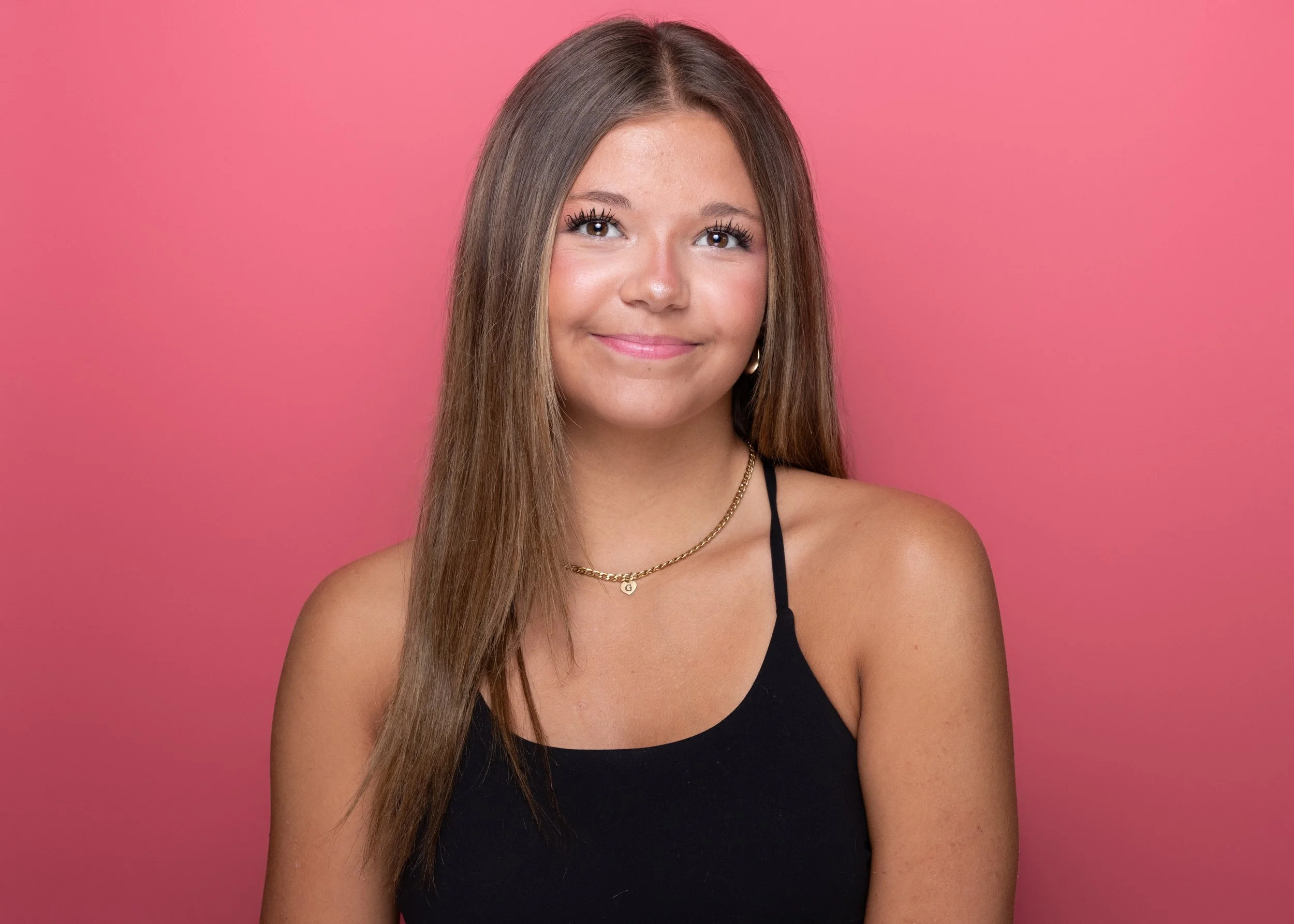 A young woman with long brown hair wearing a black spaghetti strap top, gold jewelry, and makeup, smiling against a pink background.