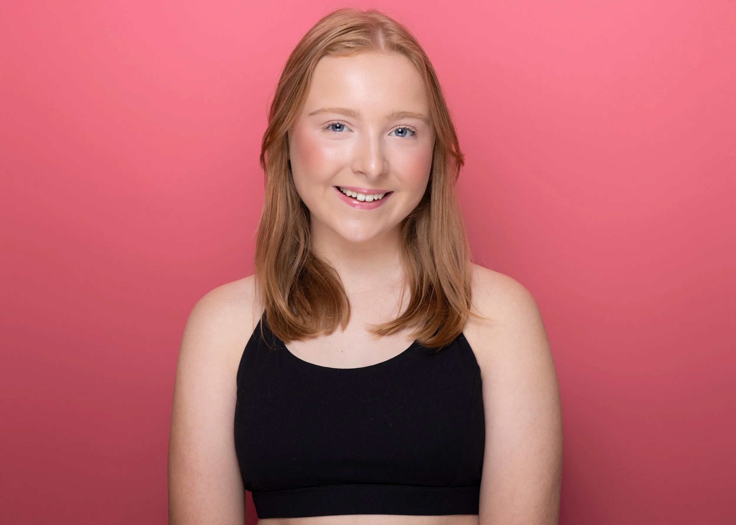 Portrait of a smiling young woman with light skin, blue eyes, and shoulder-length red hair, wearing a black sleeveless top against a pink background.
