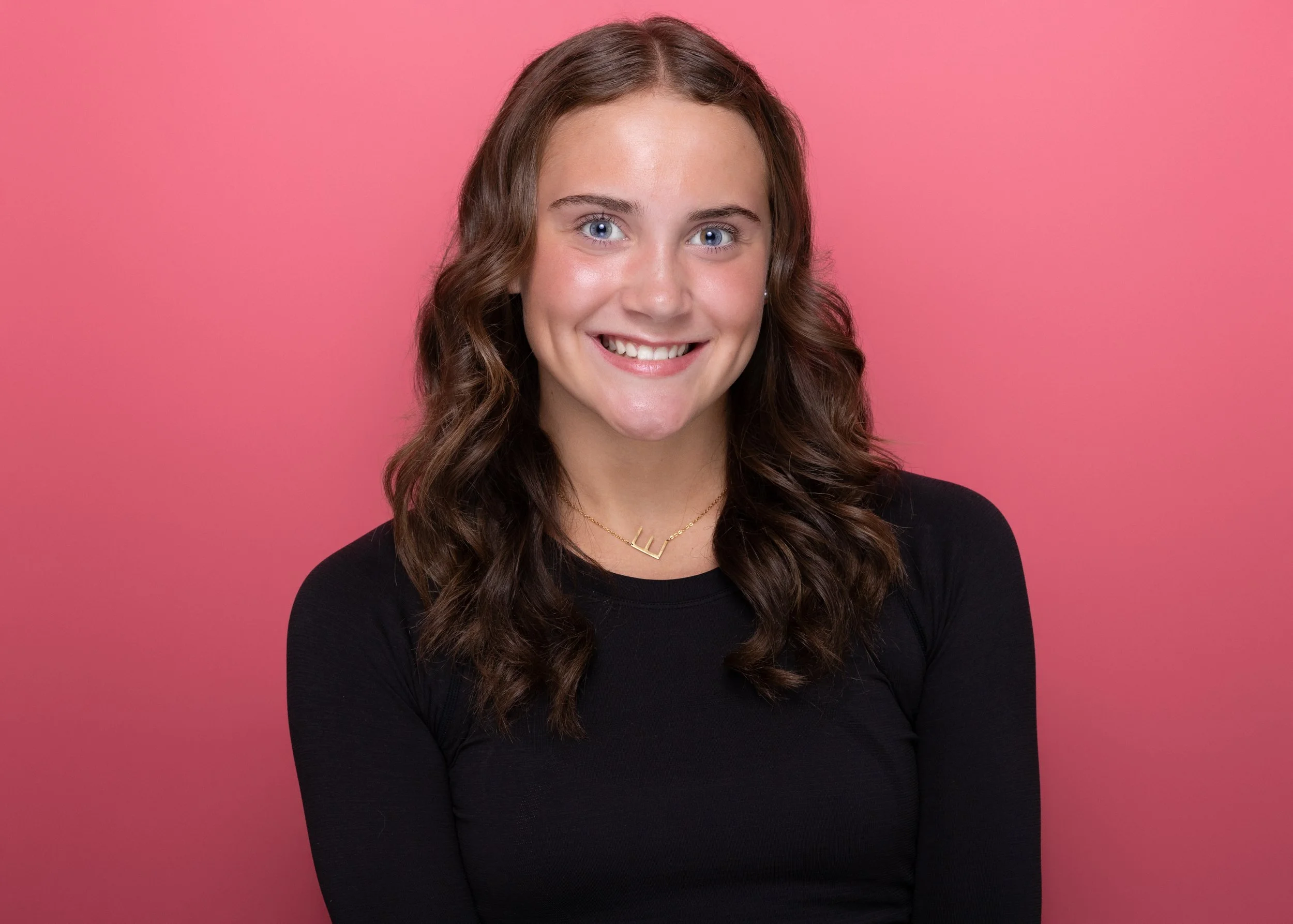 A young woman with long wavy brown hair and light skin is smiling, wearing a black long-sleeve top, a gold necklace, and pearl earrings, standing against a pink background.