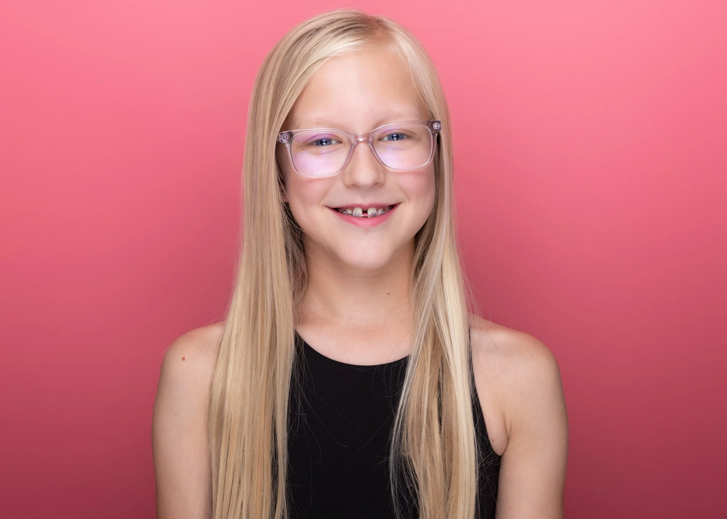 A young blonde girl wearing glasses and a black top, smiling against a pink background.