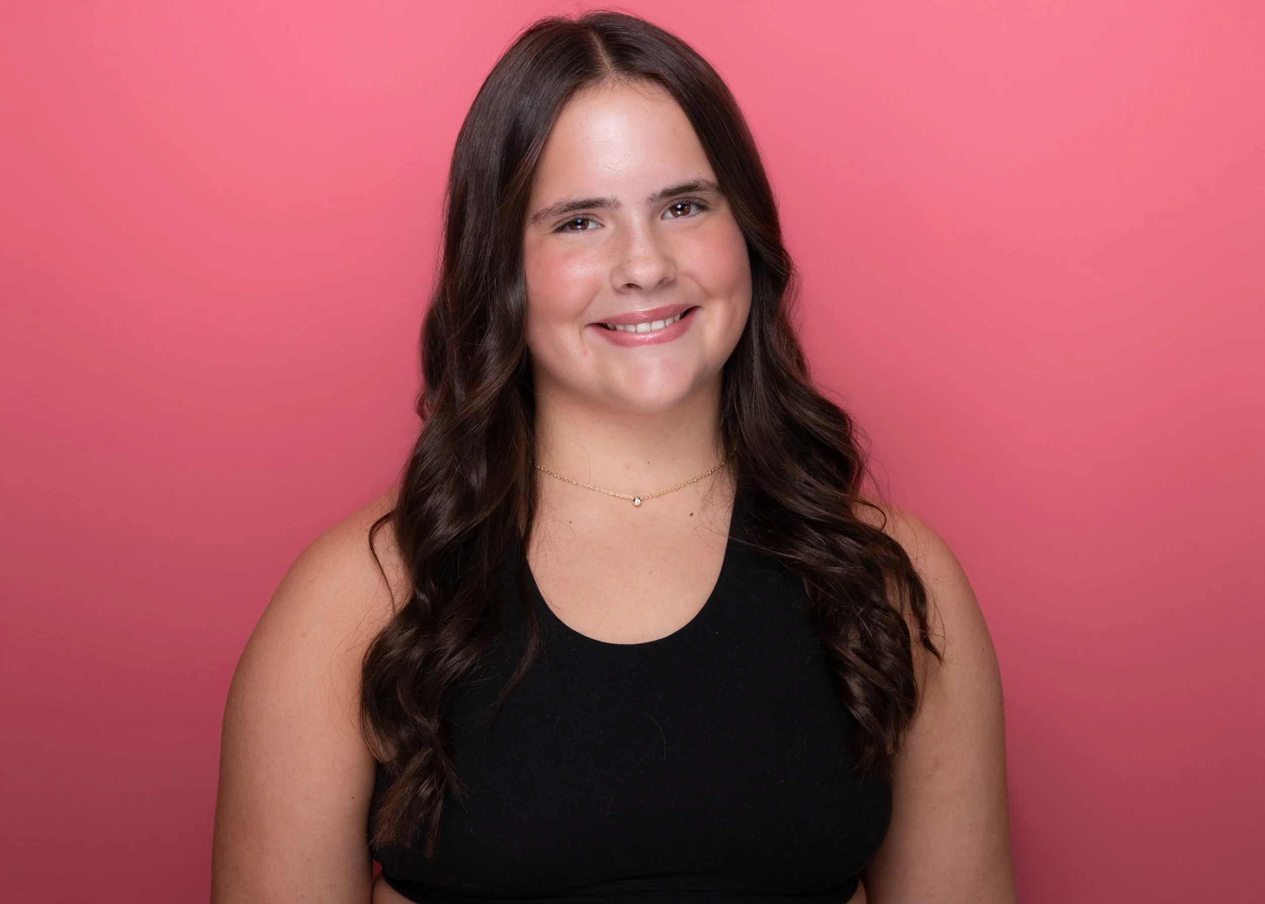 Portrait of a young woman with long wavy brown hair, smiling, wearing a black sleeveless top and a small necklace, against a pink background.