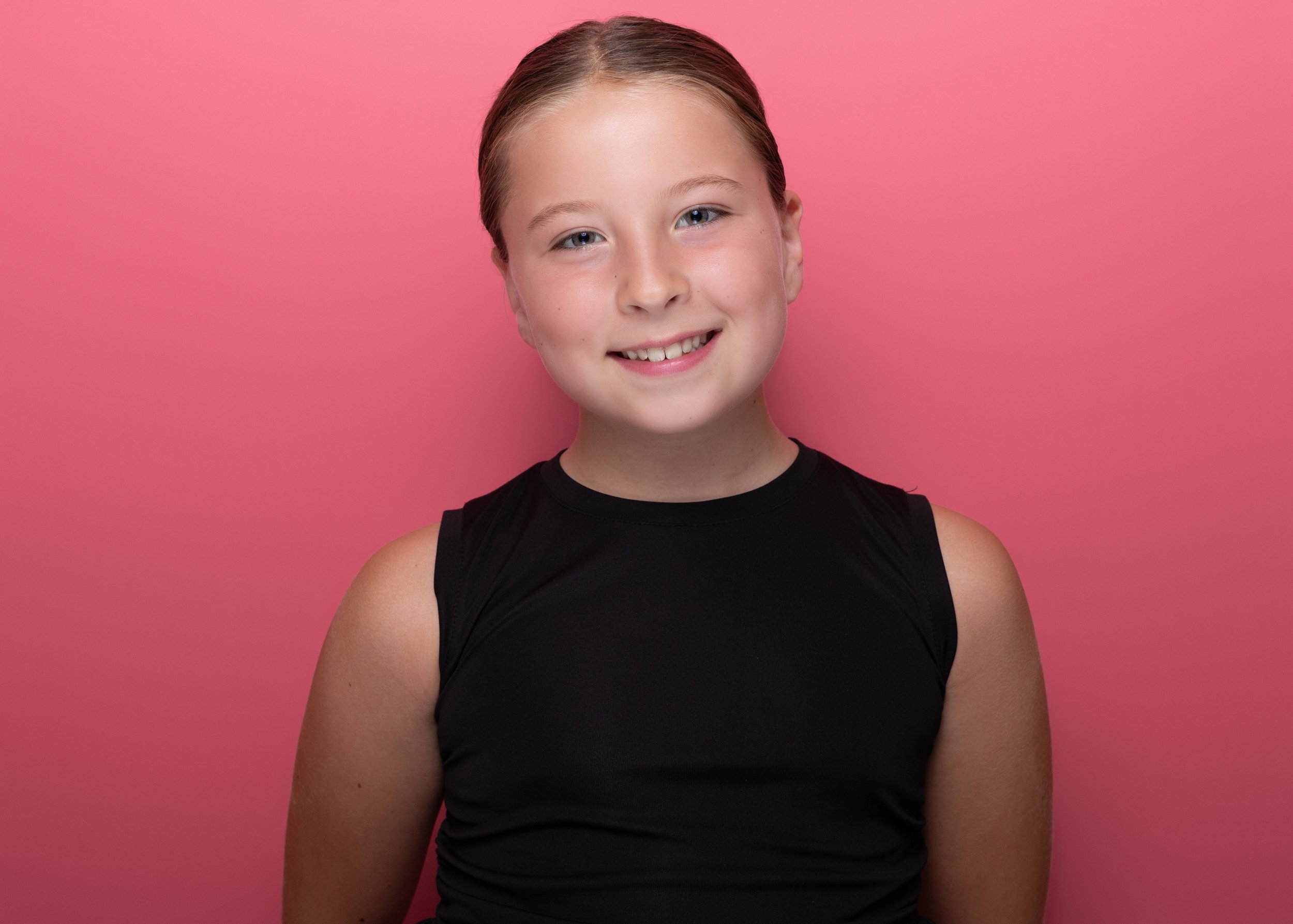 A young girl with light skin and blond hair, smiling, wearing a black sleeveless top, standing against a pink background.