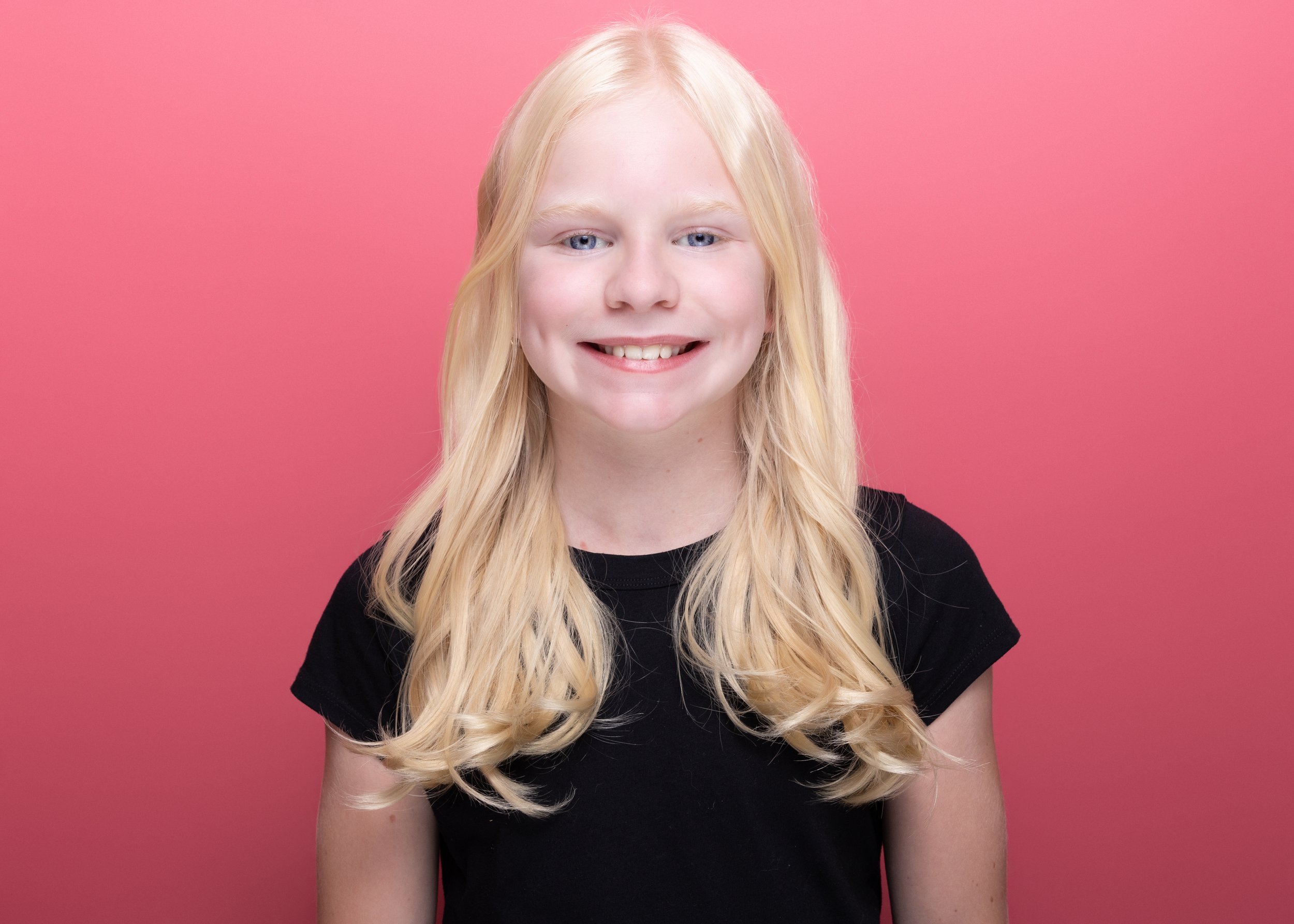 A young woman with long, wavy blonde hair, smiling, wearing a black t-shirt, standing against a pink background.