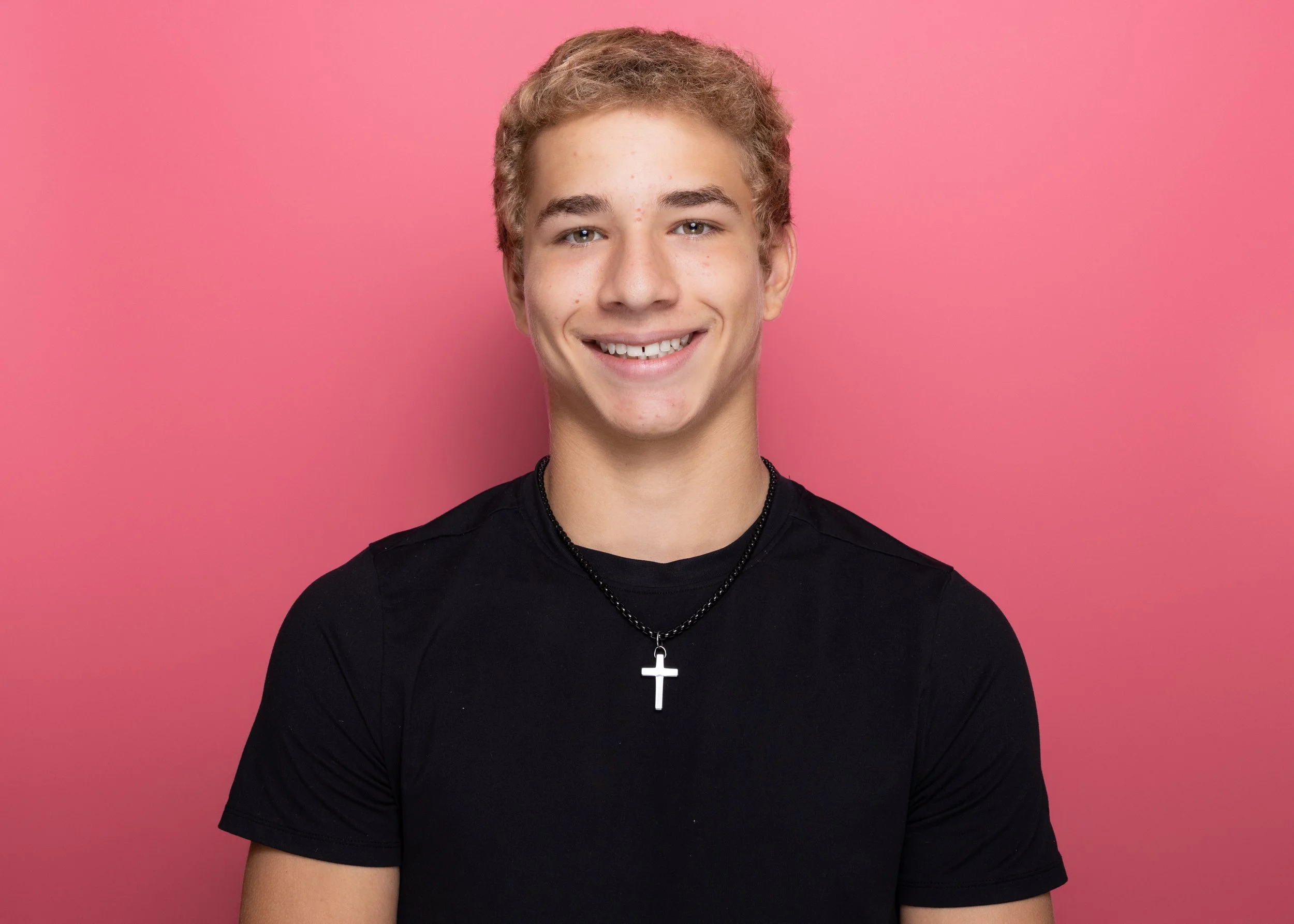 A young man with light brown curly hair and light skin, wearing a black shirt and a silver cross necklace, smiling against a pink background.