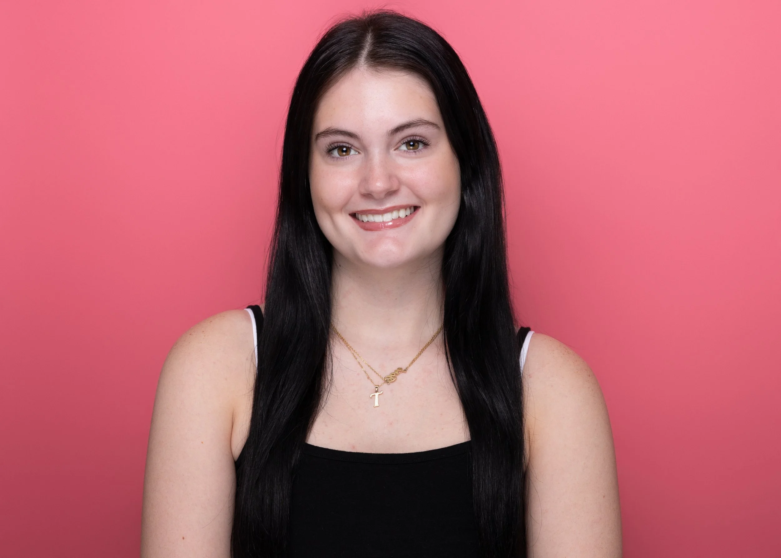 Portrait of a young woman with long dark hair, smiling, wearing a black tank top and gold necklaces, set against a pink background.