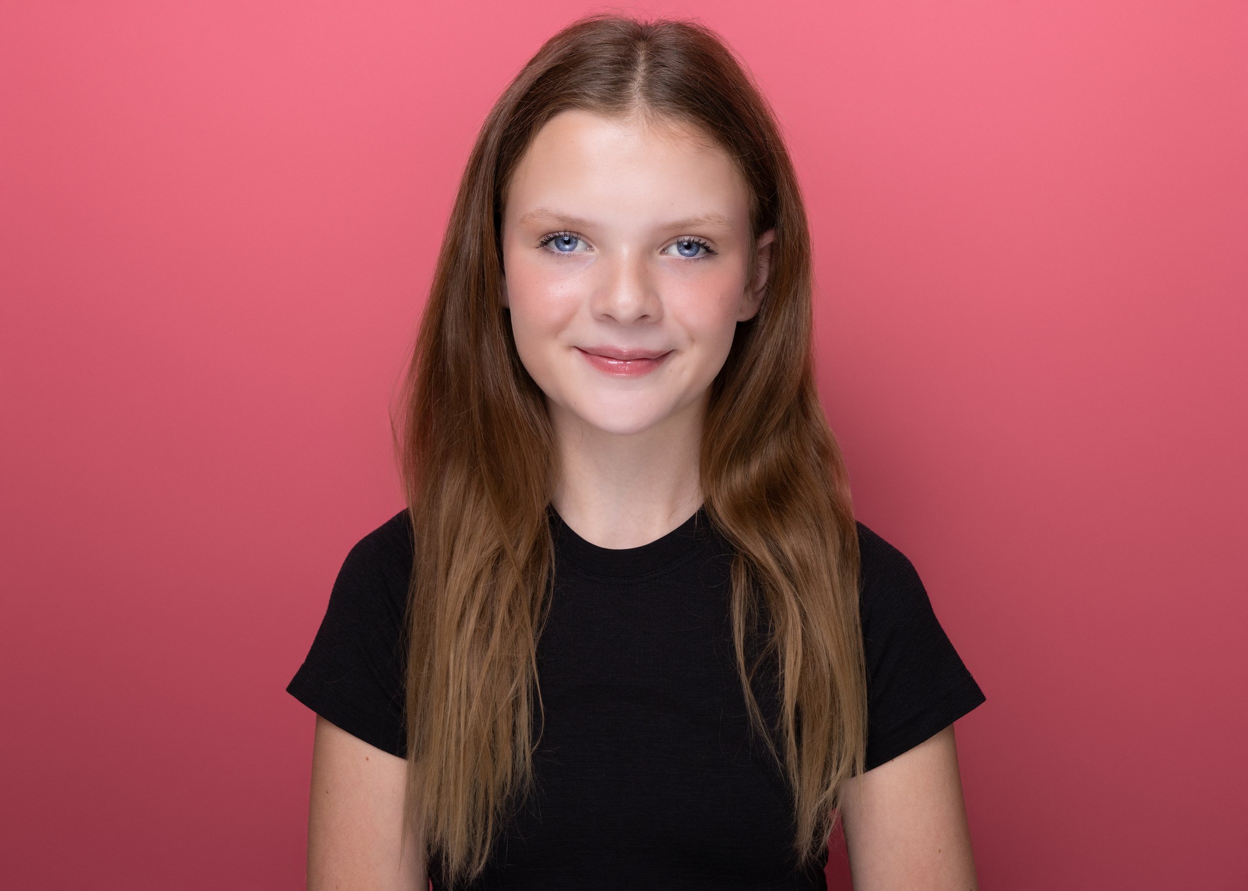 Portrait of a young woman with long brown hair, blue eyes, and light makeup, wearing a black shirt against a pink background.