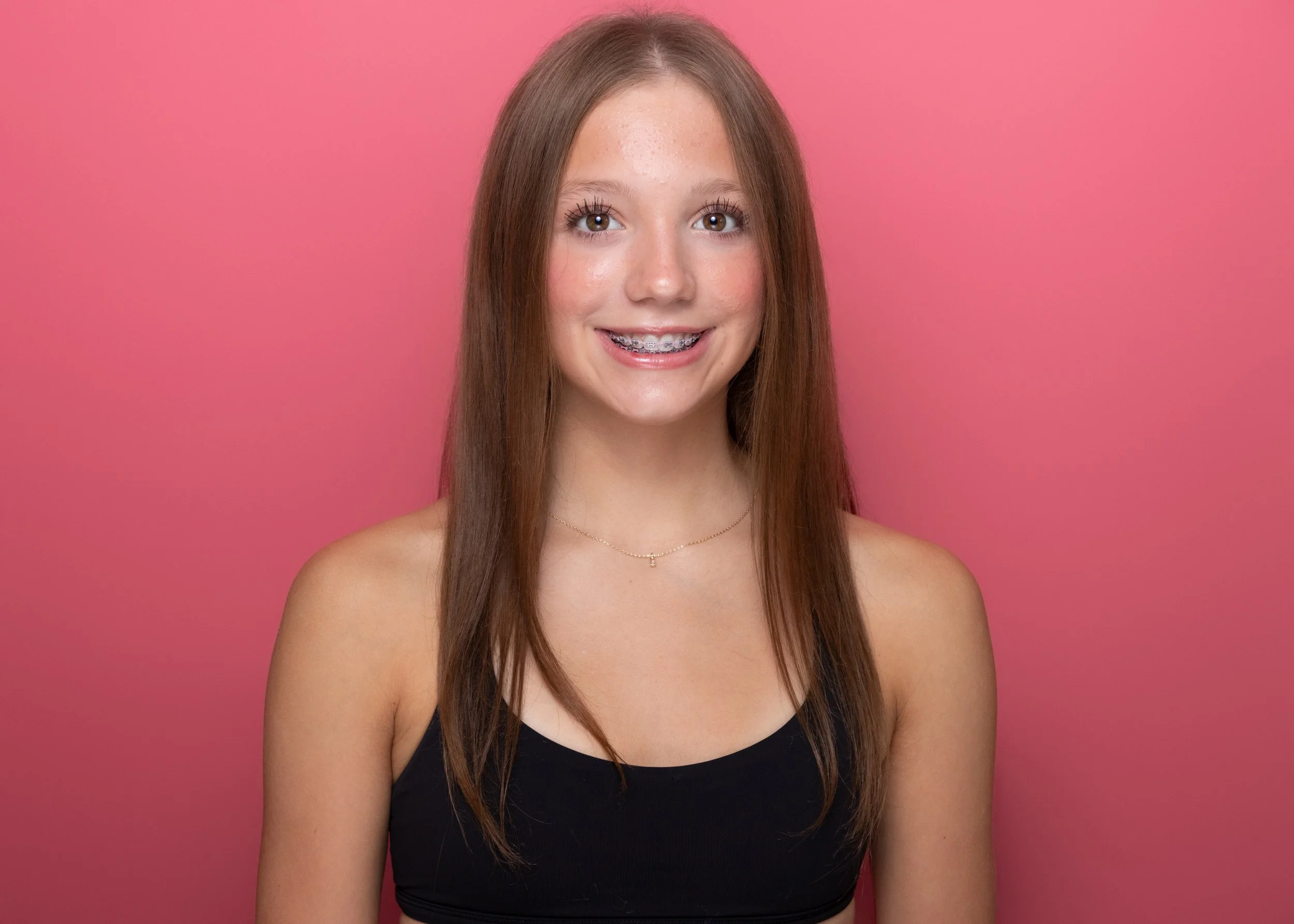 A young girl with long, straight brown hair and braces on her teeth, smiling while wearing a black tank top and a delicate necklace, standing against a pink background.