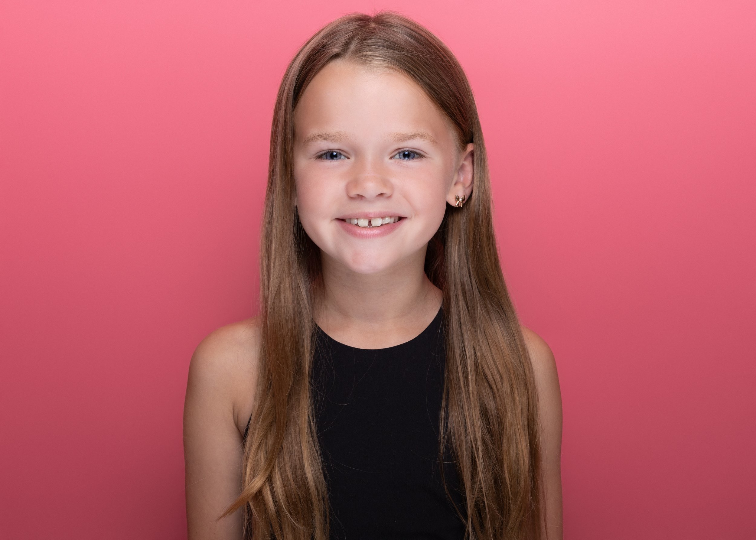 Portrait of a young girl with long red hair, blue eyes, and a smile, wearing a black sleeveless top, against a pink background.