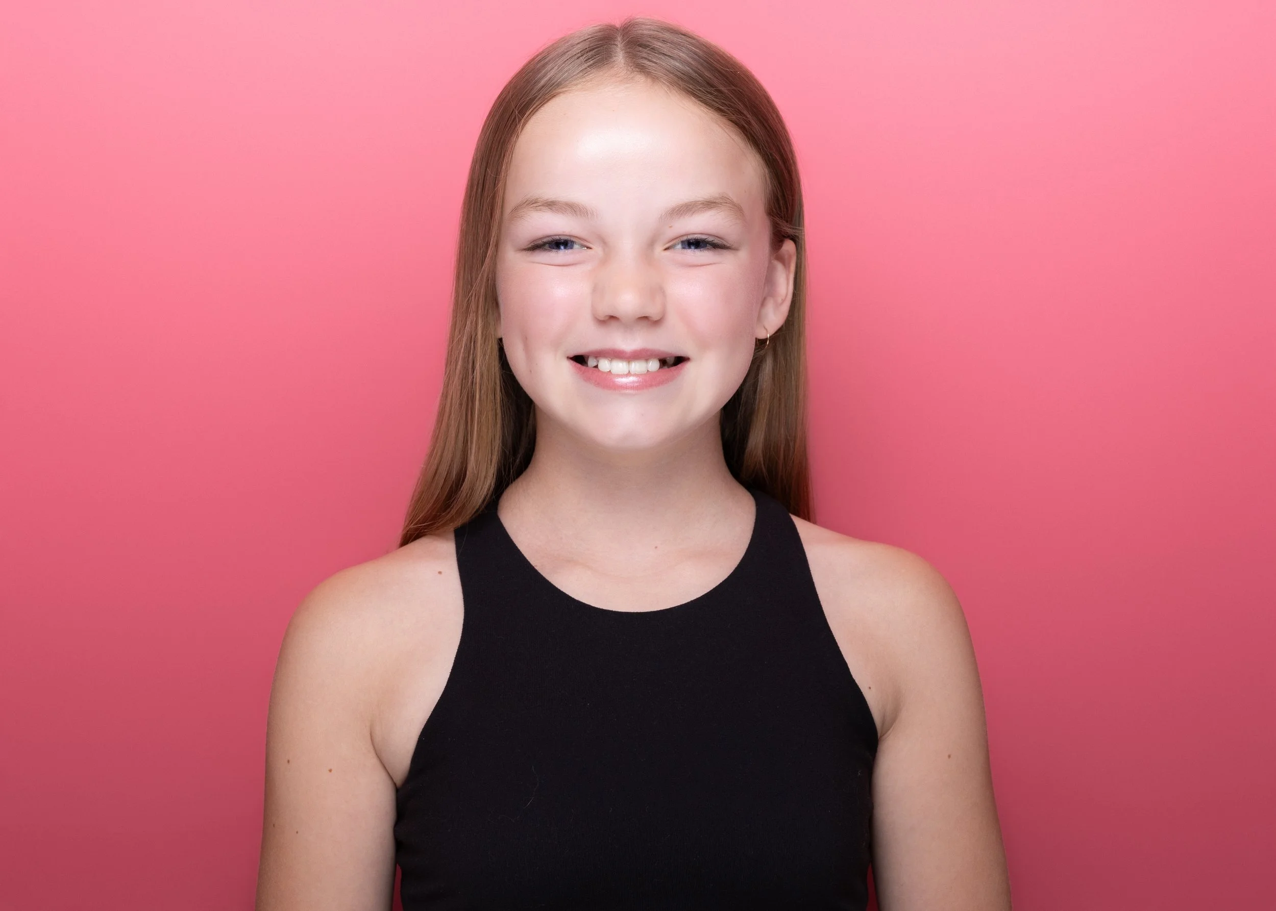 A young woman with long red hair, light skin, and a bright smile, wearing a sleeveless black top, stands against a pink background.