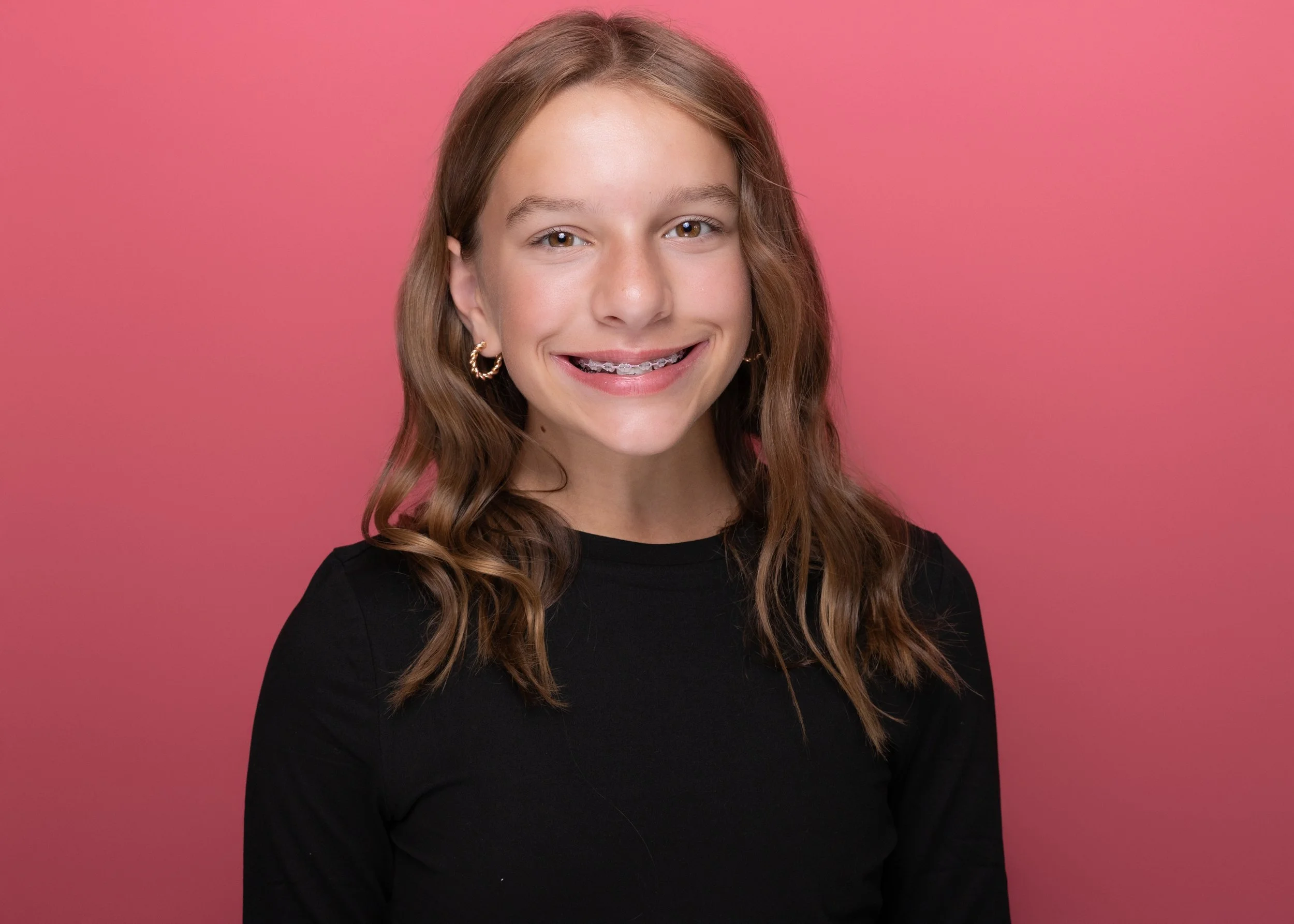 Close-up portrait of a young woman with wavy brown hair, smiling with braces, wearing gold hoop earrings and a black top, against a pink background.