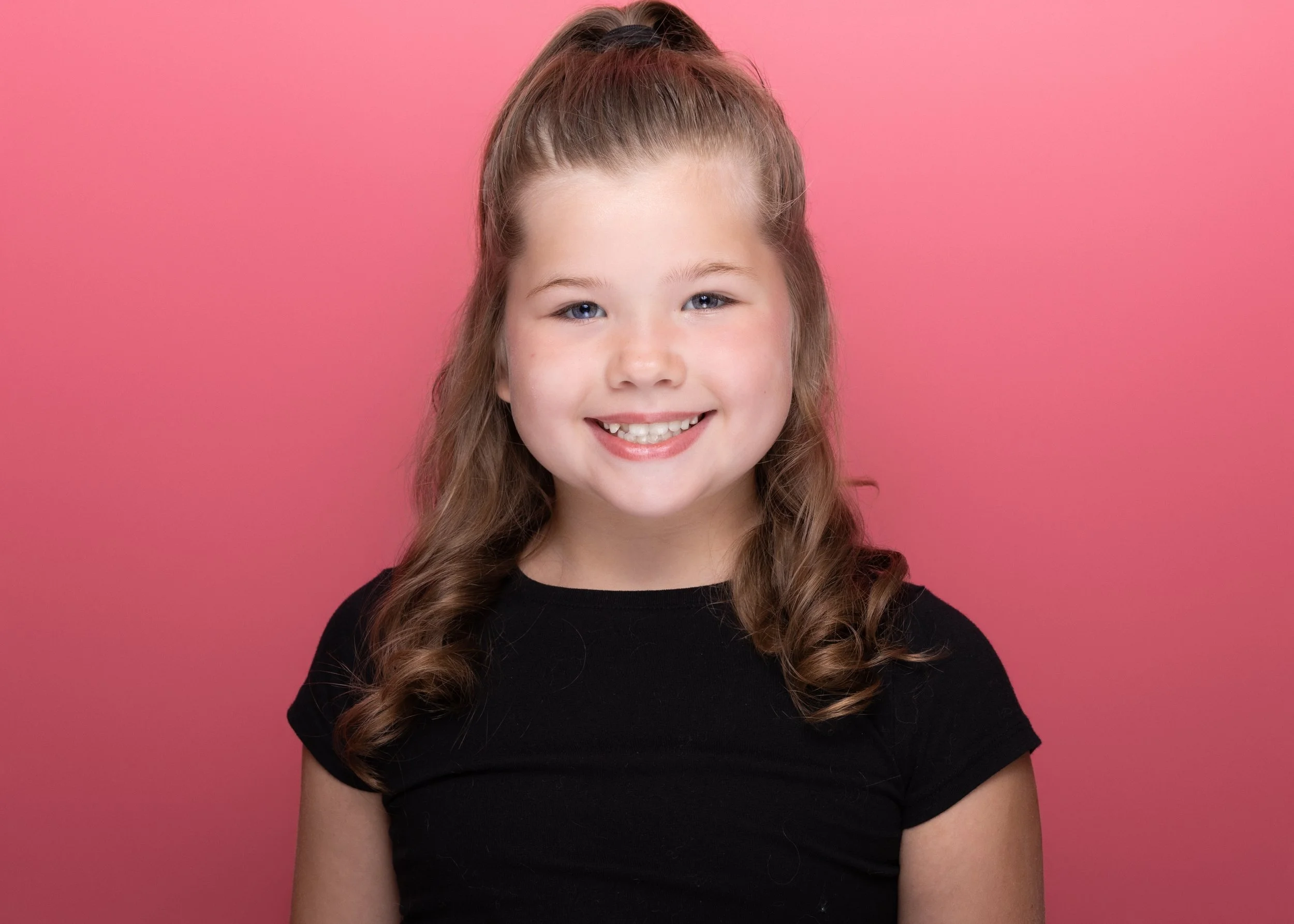 A young girl with long wavy hair smiling in front of a pink background.