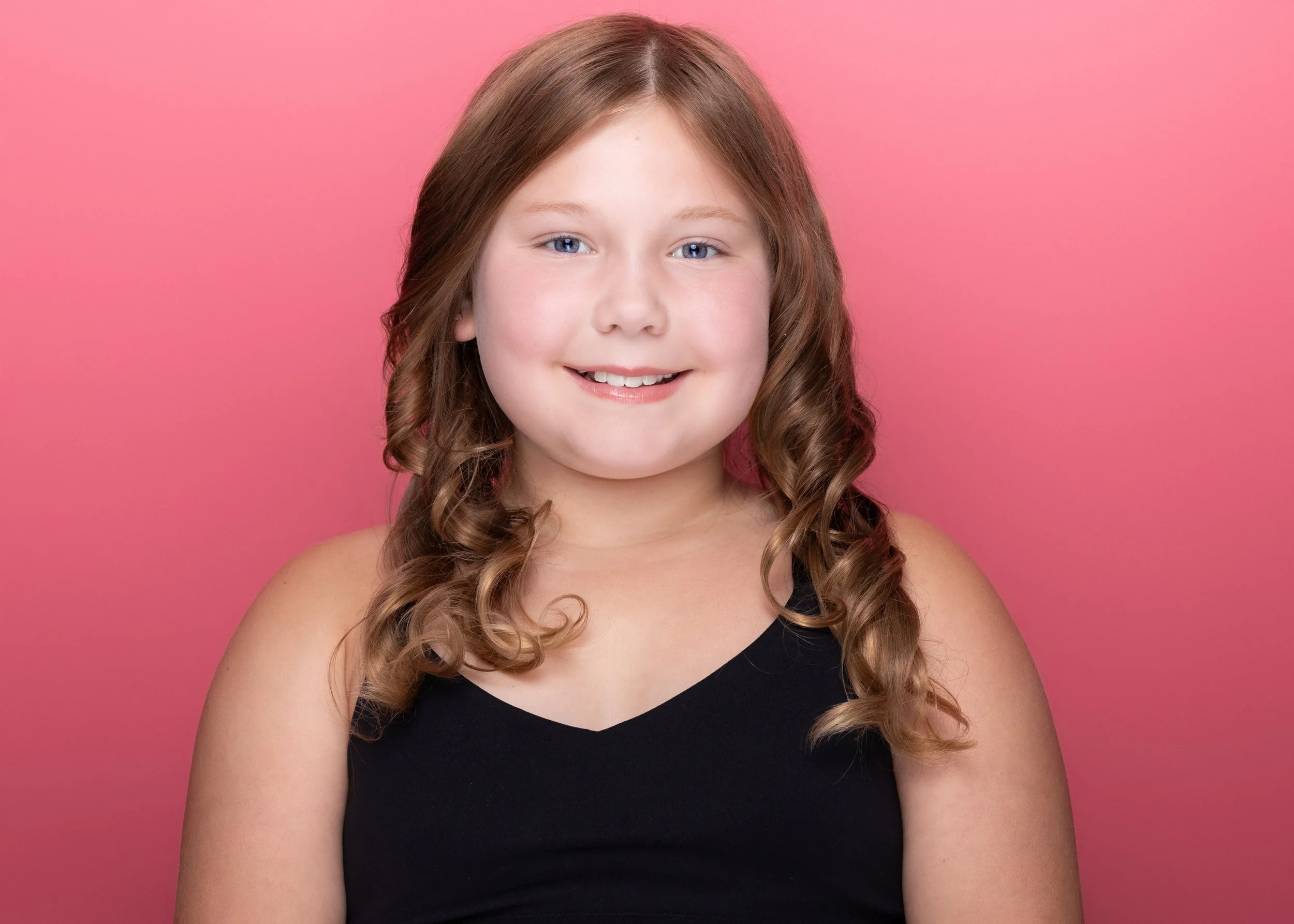 A young girl with curly light brown hair and blue eyes, wearing a black tank top, smiling against a pink background.