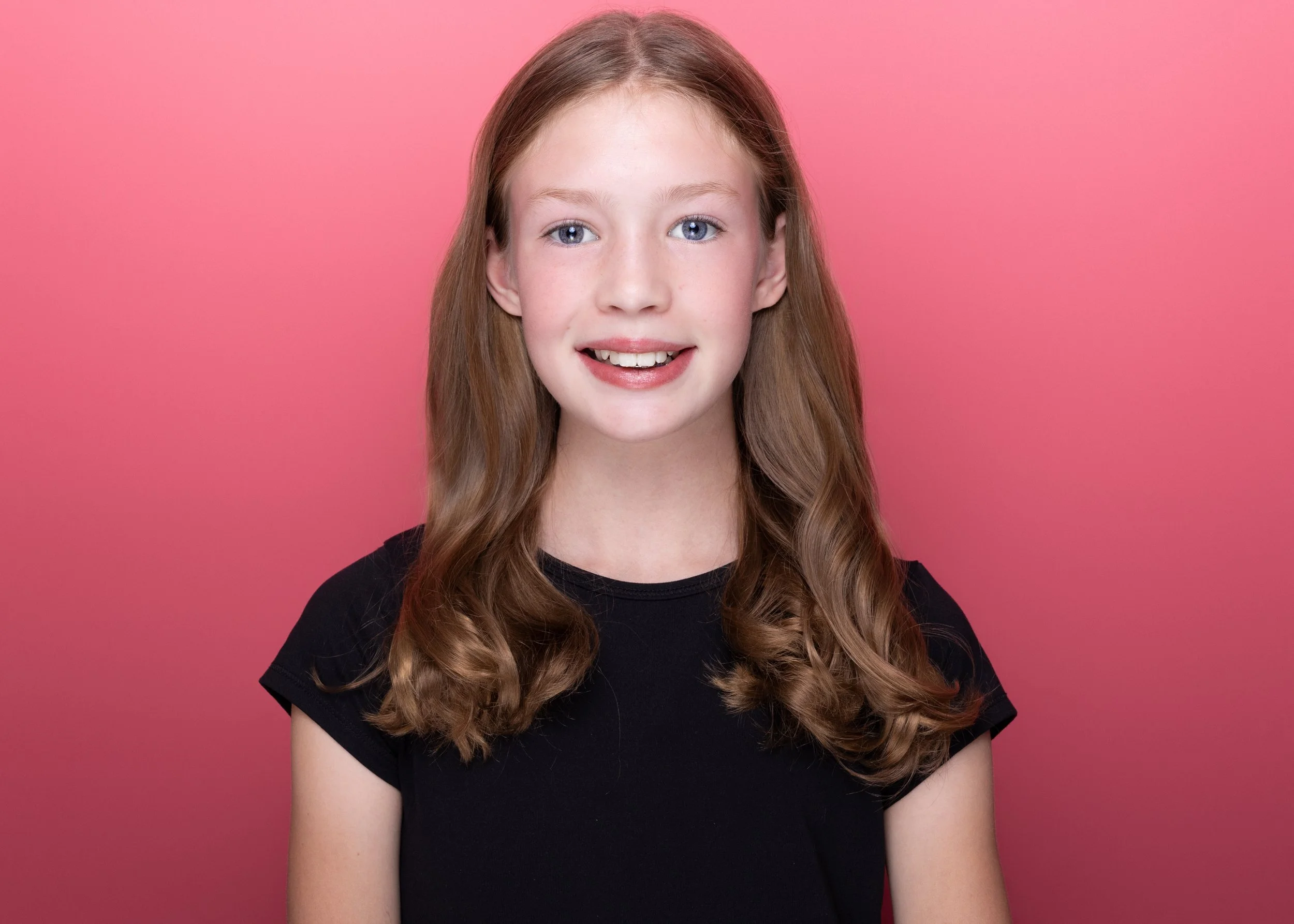Portrait of a young girl with long, wavy red hair, blue eyes, freckles, wearing a black t-shirt, smiling, against a pink background.
