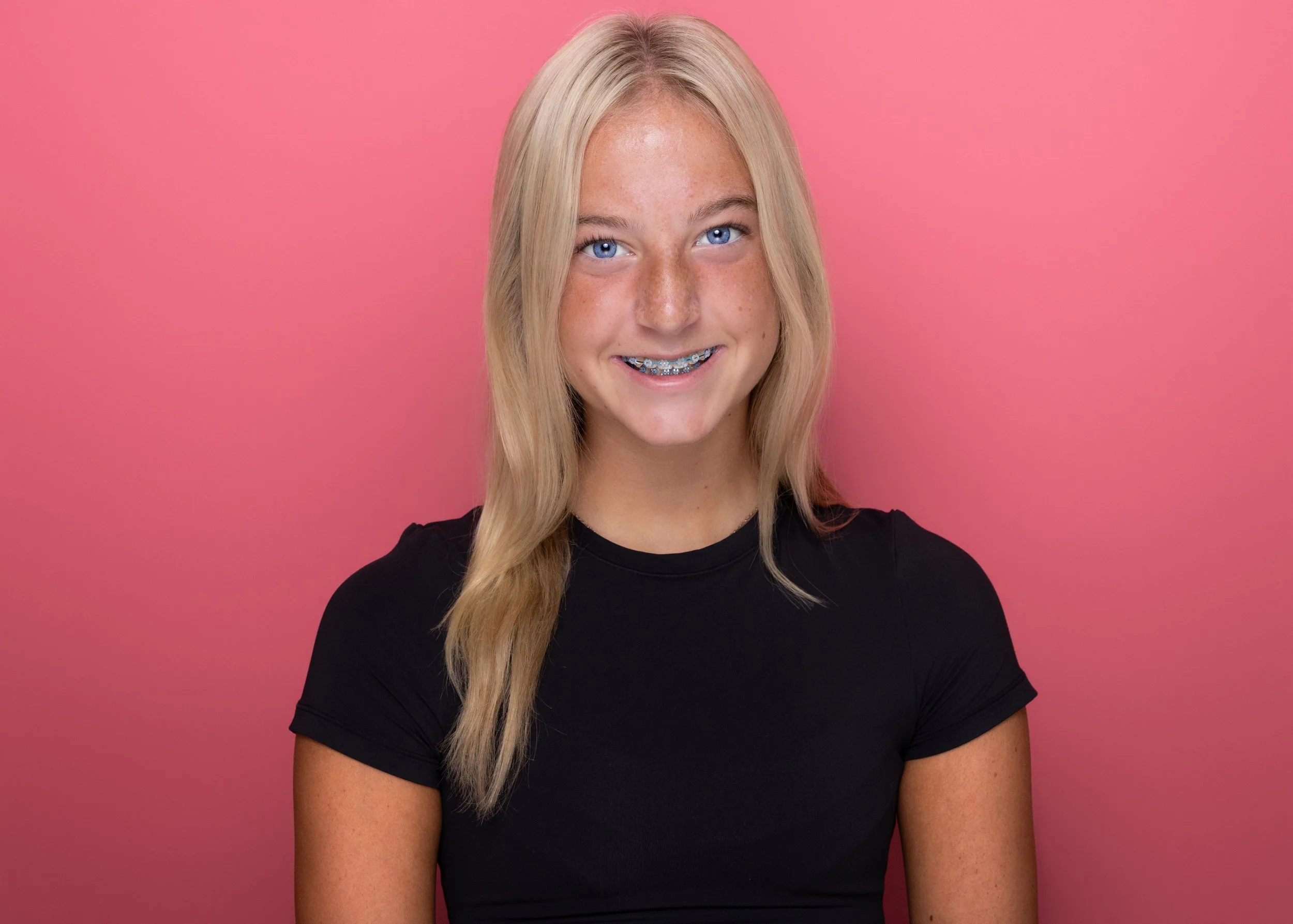 A young girl with long blonde hair, blue eyes, and braces on her teeth, smiling, wearing a black shirt, against a pink background.
