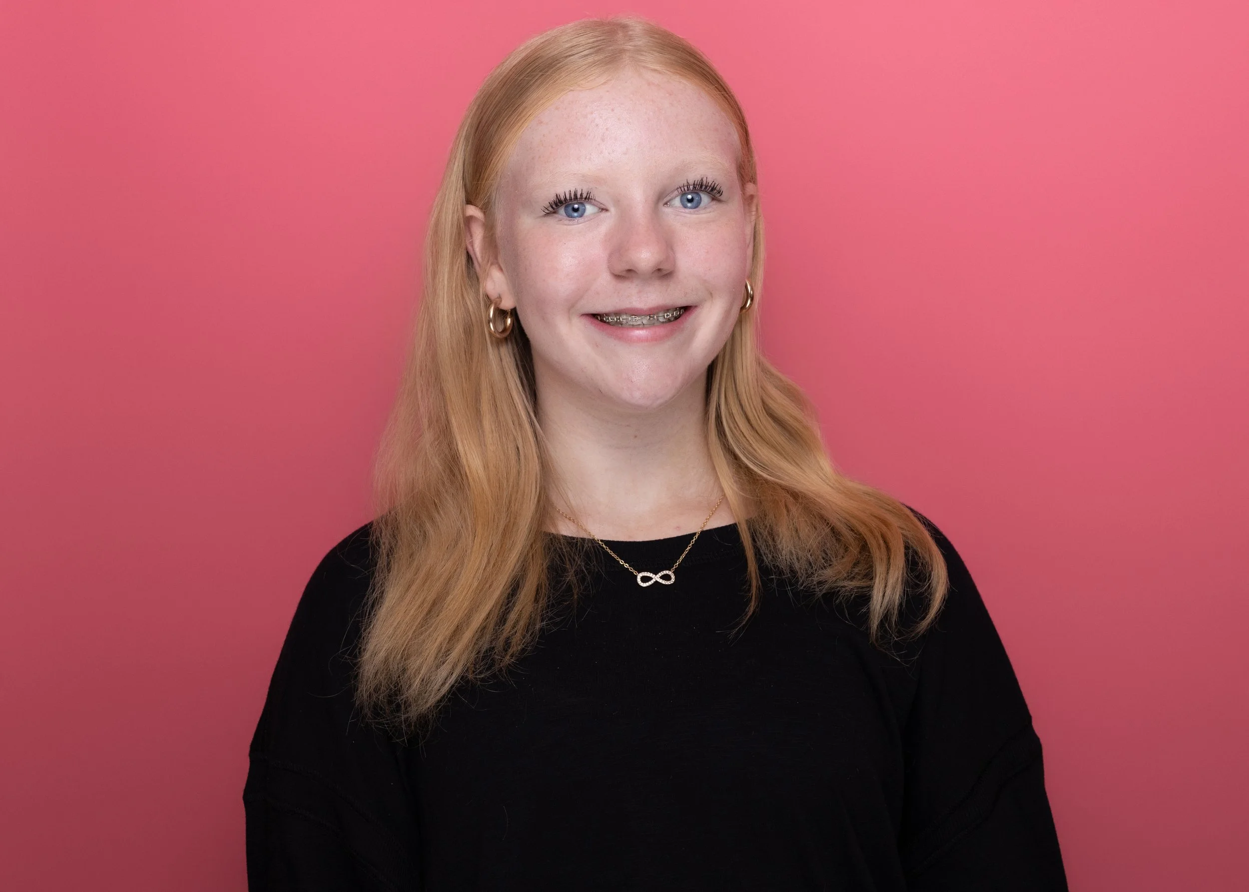 A young girl with long red hair, wearing a black top, gold hoop earrings, a necklace with an infinity symbol, and braces, smiling in front of a pink background.