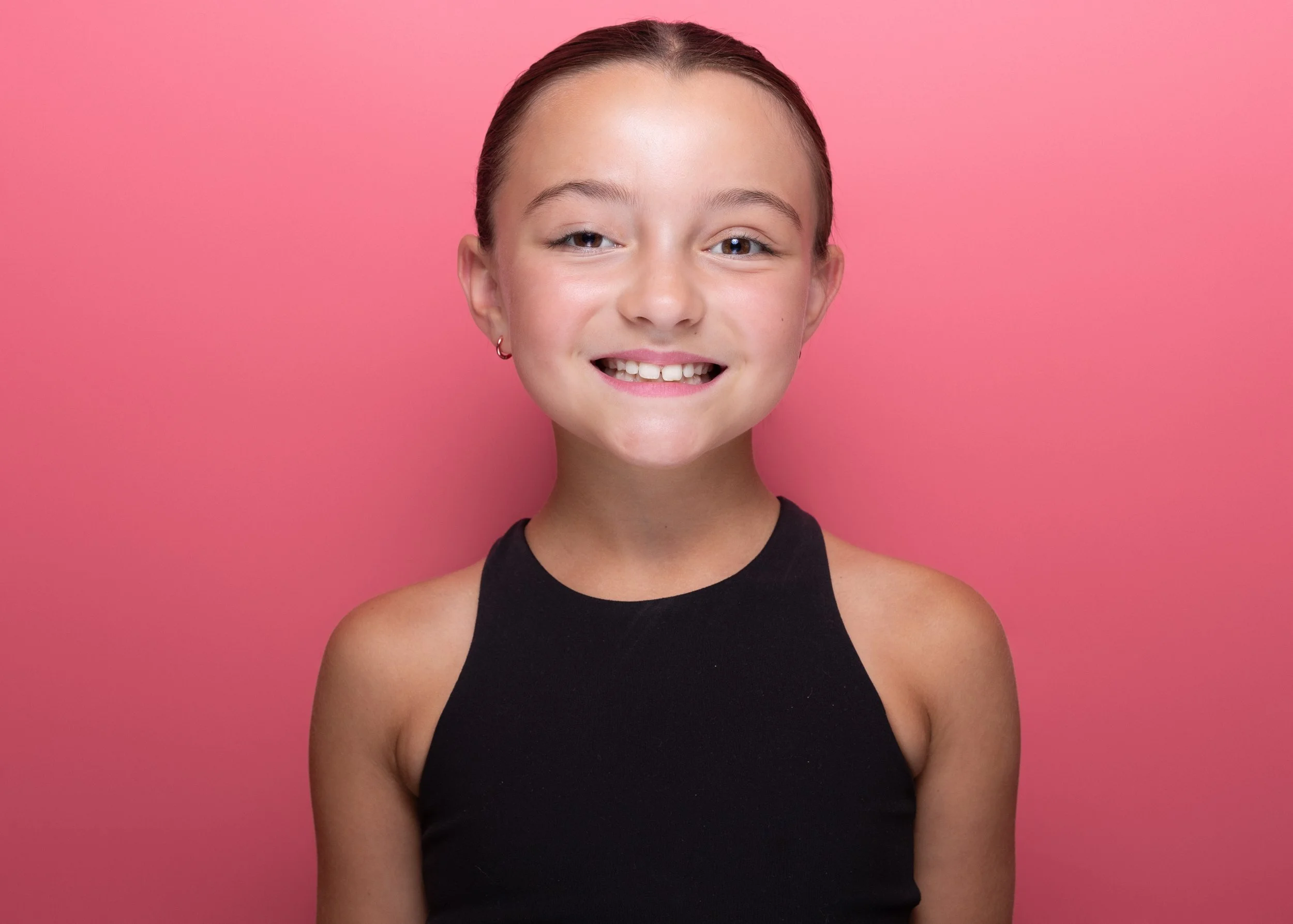 A young woman with a short, tied-back hairstyle, wearing hoop earrings and a black sleeveless top, smiling against a pink background.