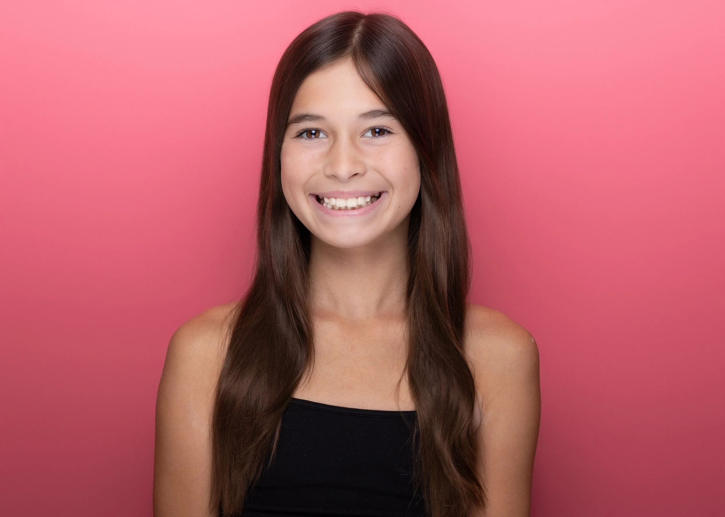 A young girl with long brown hair, smiling, wearing a black top, against a pink background.