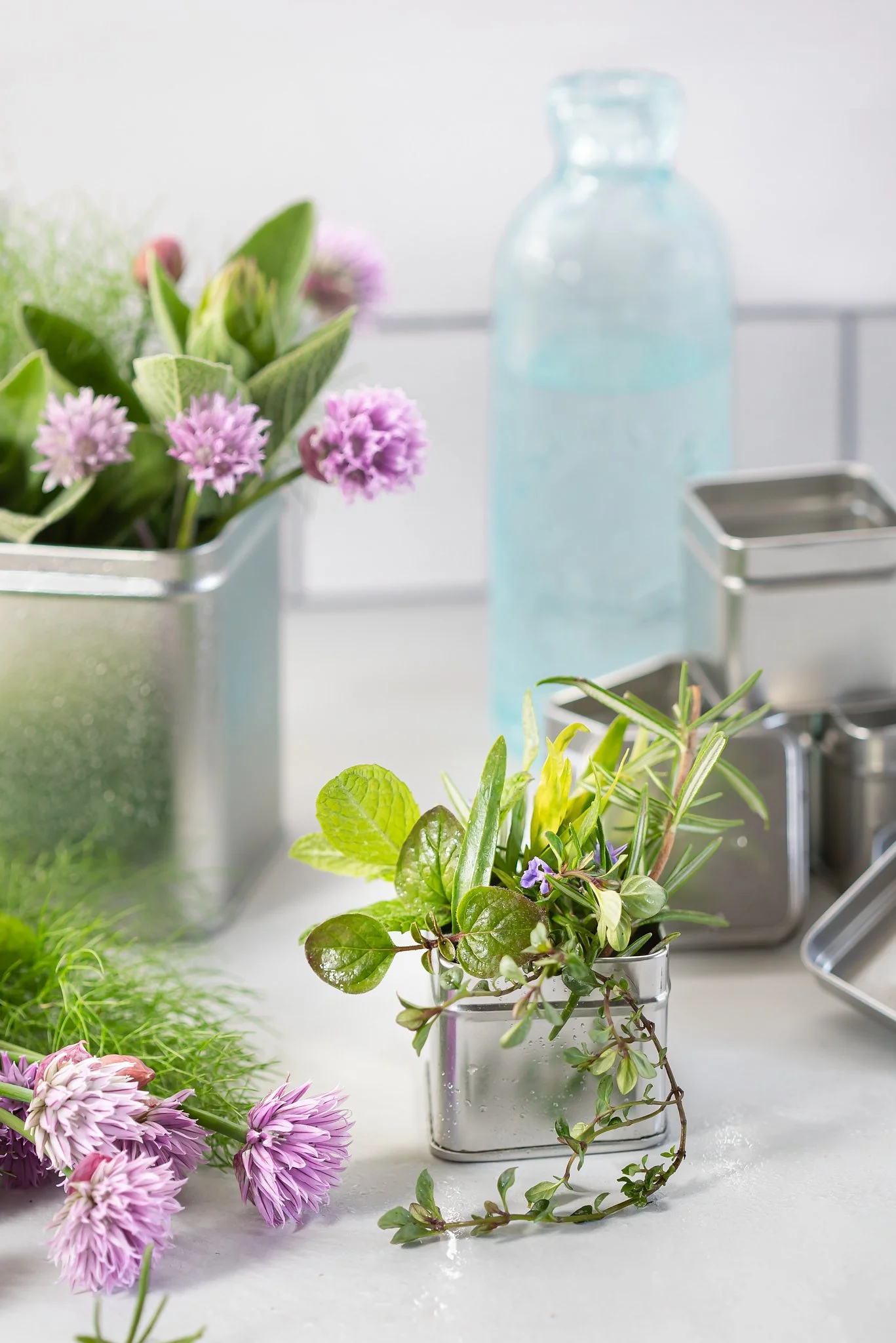 A collection of fresh green herbs and purple flowers on a white kitchen countertop, with metal containers and a glass water bottle in the background.