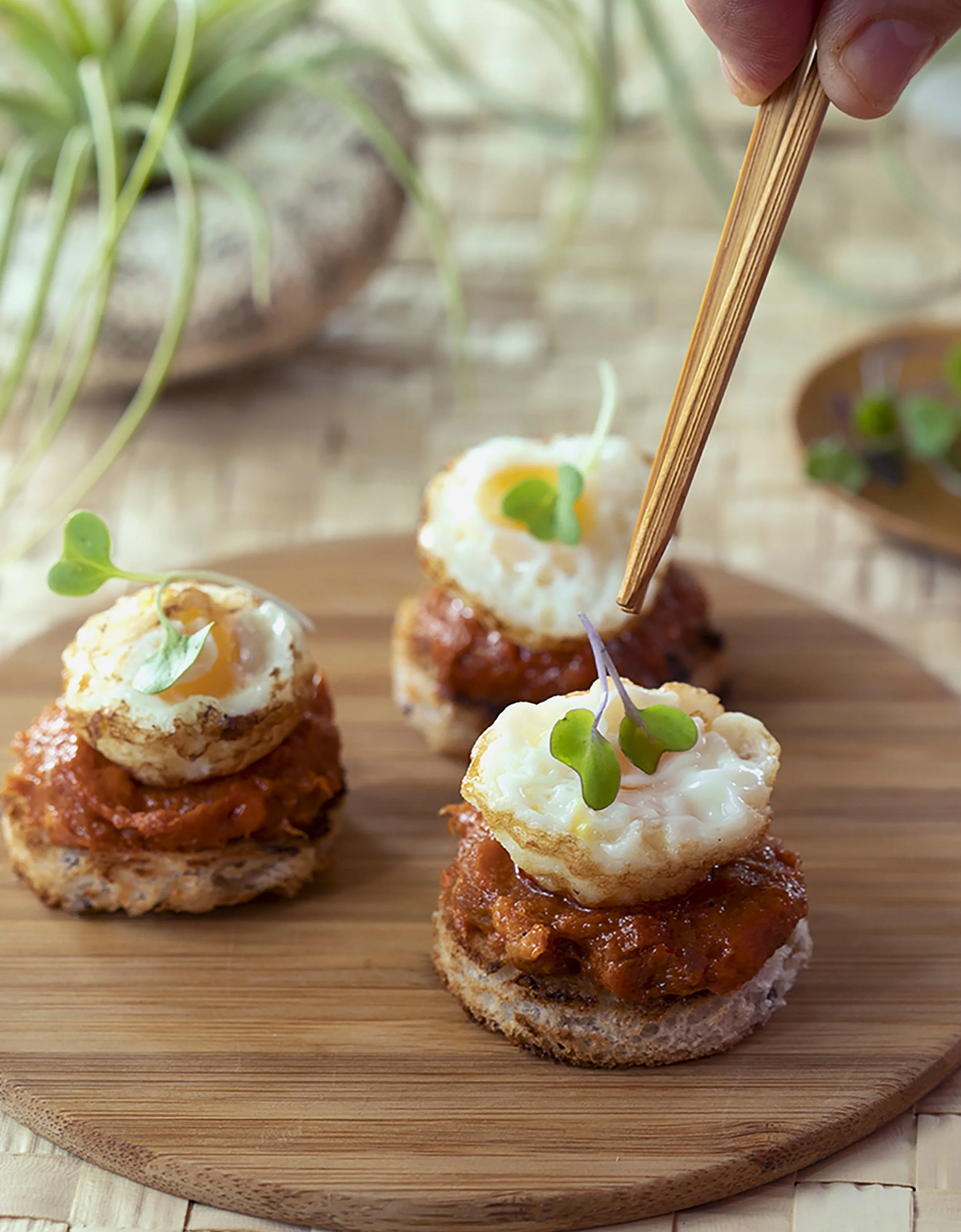 Appetizers with small bread pieces topped with spicy meat sauce, a dollop of sour cream, and microgreens on a round wooden serving board.