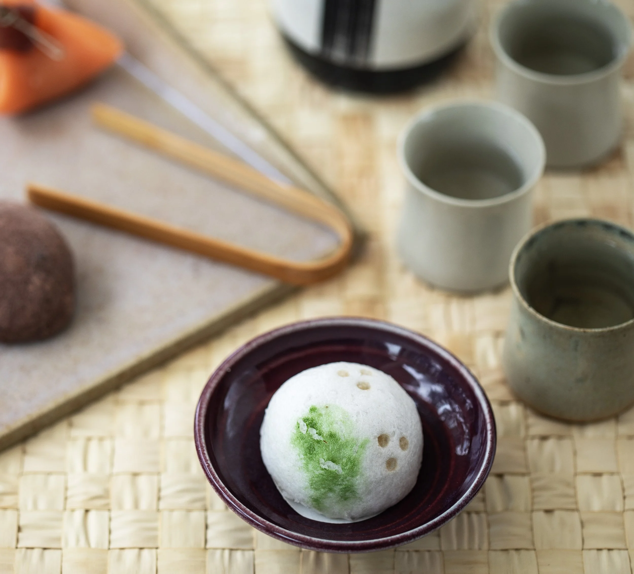 A traditional Japanese tea set with three ceramic cups, a plate with a white dessert featuring green decoration, bamboo utensils, and a textured mat.