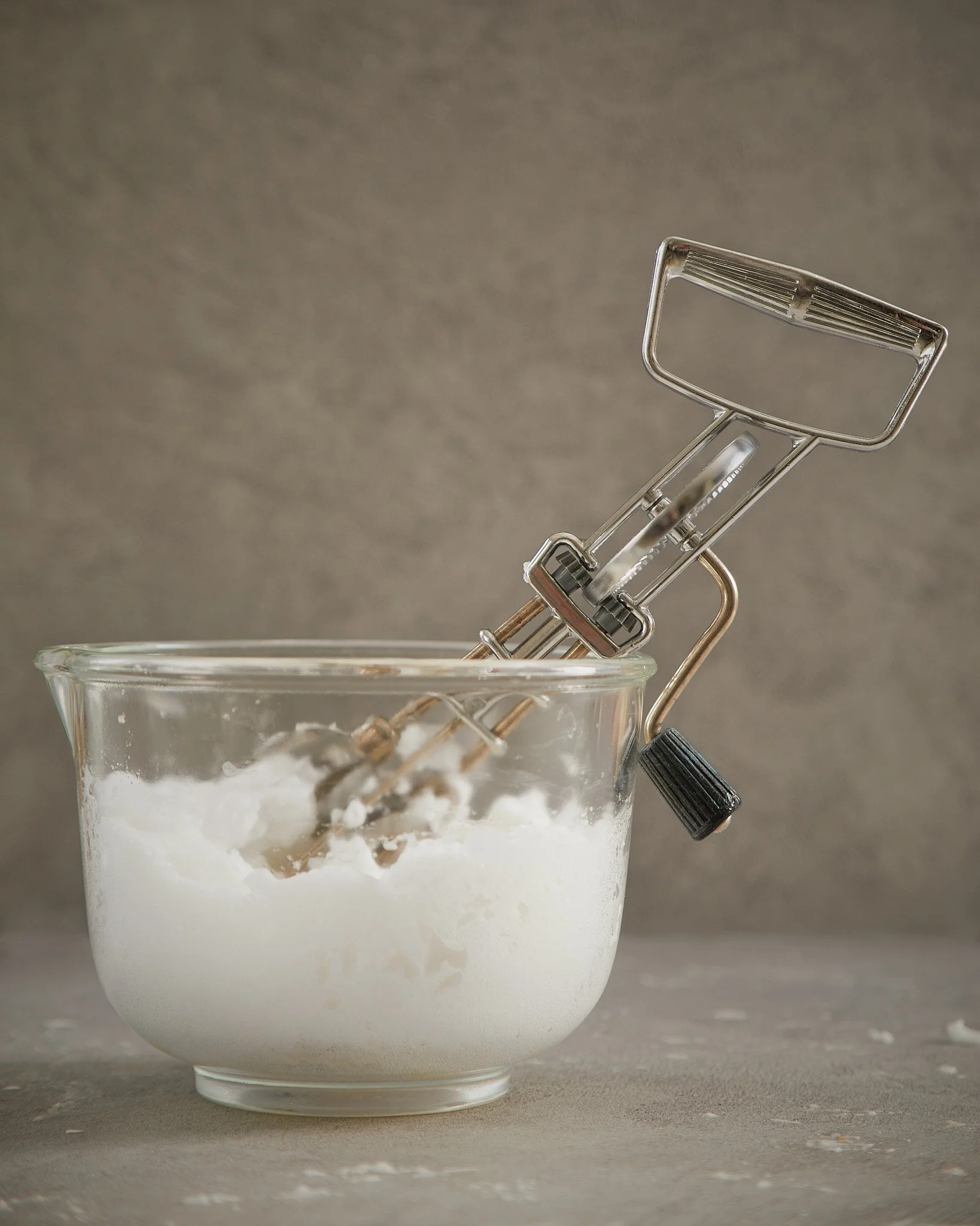 A glass mixing bowl with flour and a hand mixer inside, on a gray surface with a neutral background.