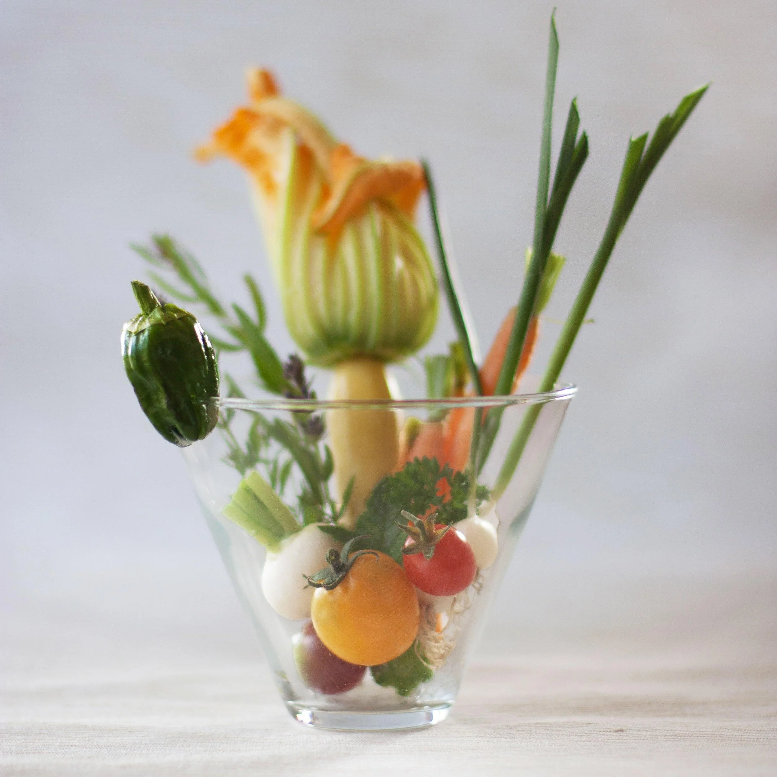 A glass bowl filled with various fresh vegetables, including cherry tomatoes, green onion, celery, carrot, broccoli, and chives on a light surface with a plain background.