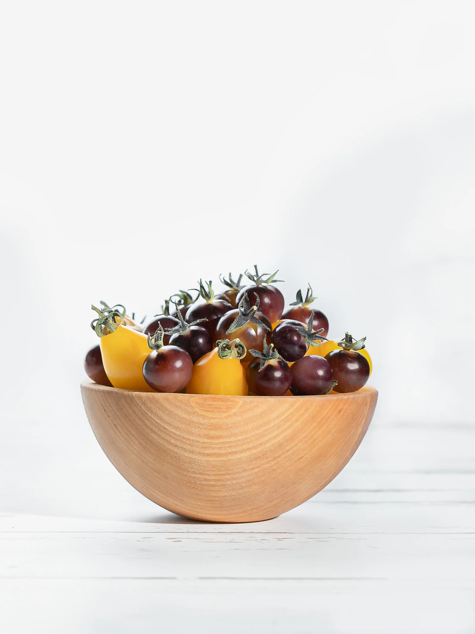 A wooden bowl filled with yellow and purple cherry tomatoes on a white surface against a white background.