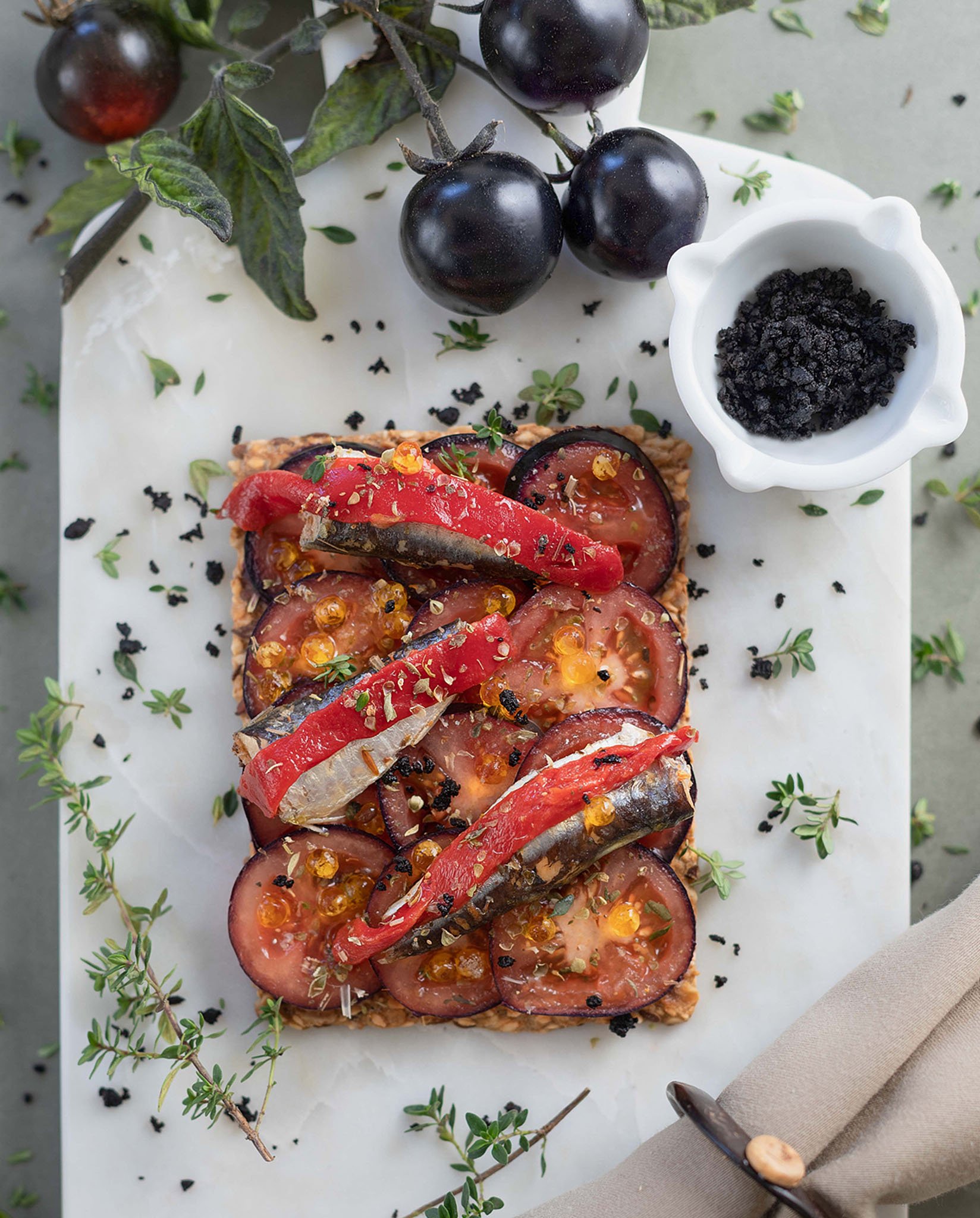Open-faced tomato and eggplant tart garnished with herbs, black salt, and pepper, with fresh black grapes and a small bowl of black salt nearby.
