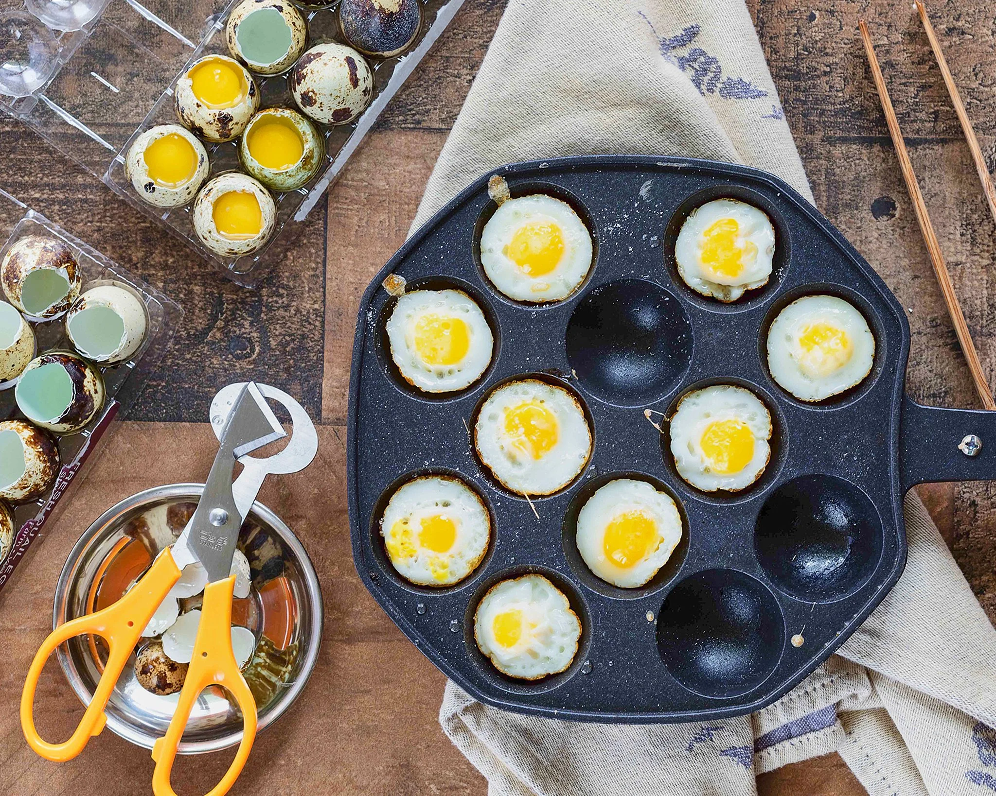 A black takoyaki maker with several cooked eggs inside and empty slots, on a wooden surface with pair of chopsticks, a cloth, and eggs in plastic trays nearby.