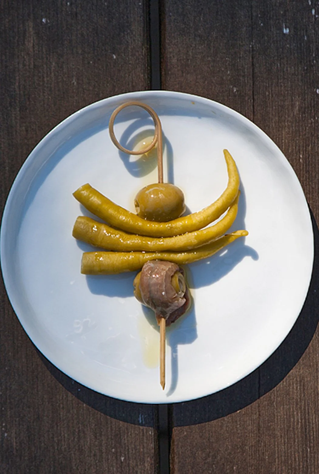 A white plate with green olives, pickled peppers, and a piece of cured meat on a skewer, set on a wooden table.