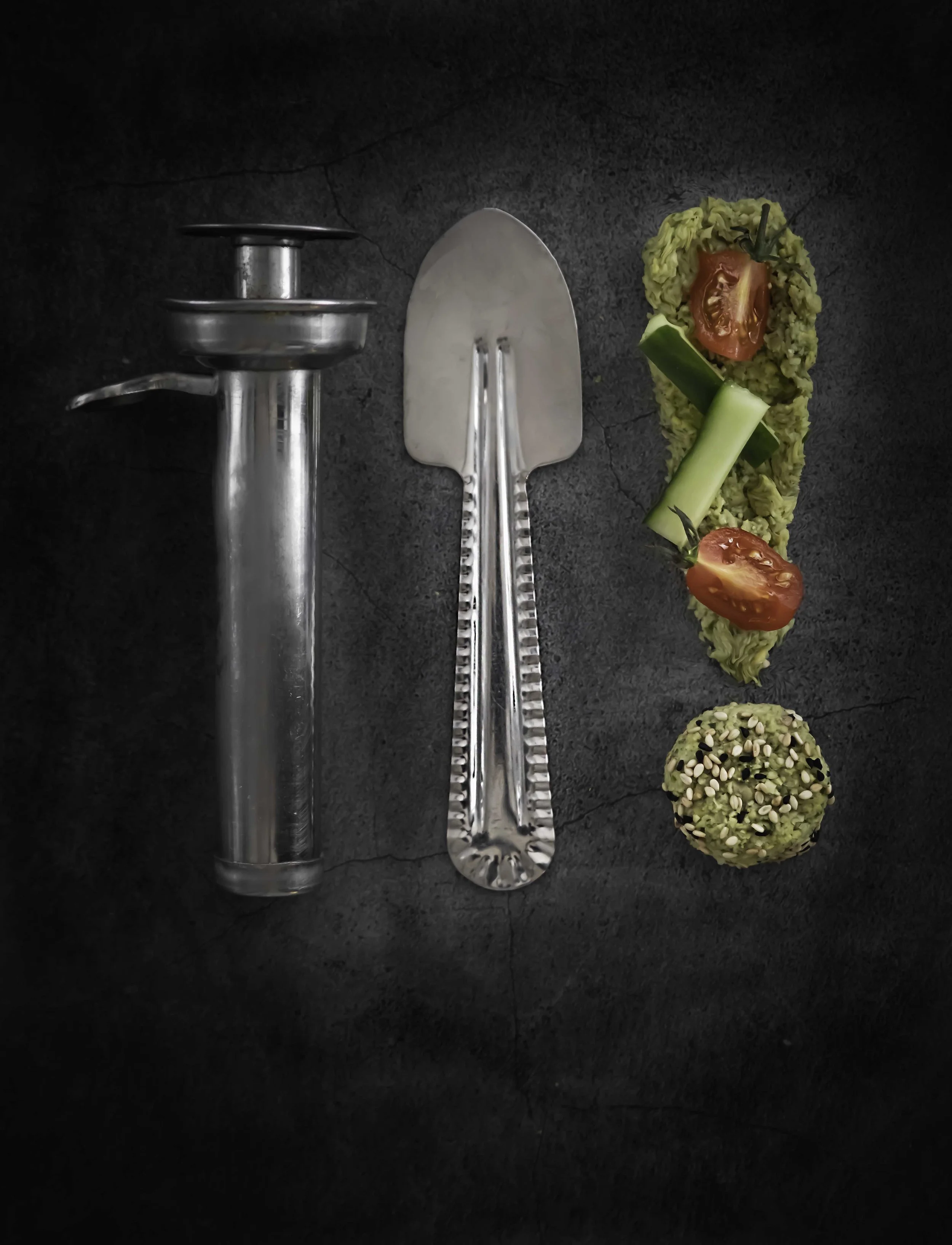 Flat lay of a stainless steel garlic press and a metal scoop with a black textured background; beside them, there's a piece of sushi with green filling, cherry tomato, cucumber, and sesame seed ball.