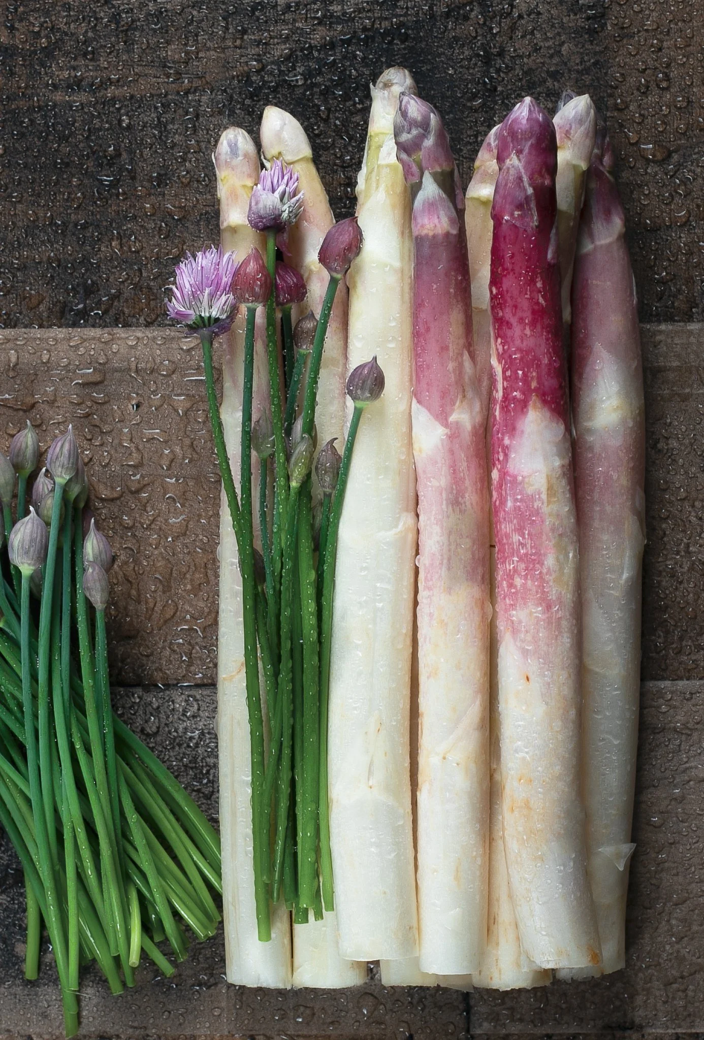 Fresh white and pink asparagus stalks, with some purple flower buds and blossoms, resting on a wet wooden surface.