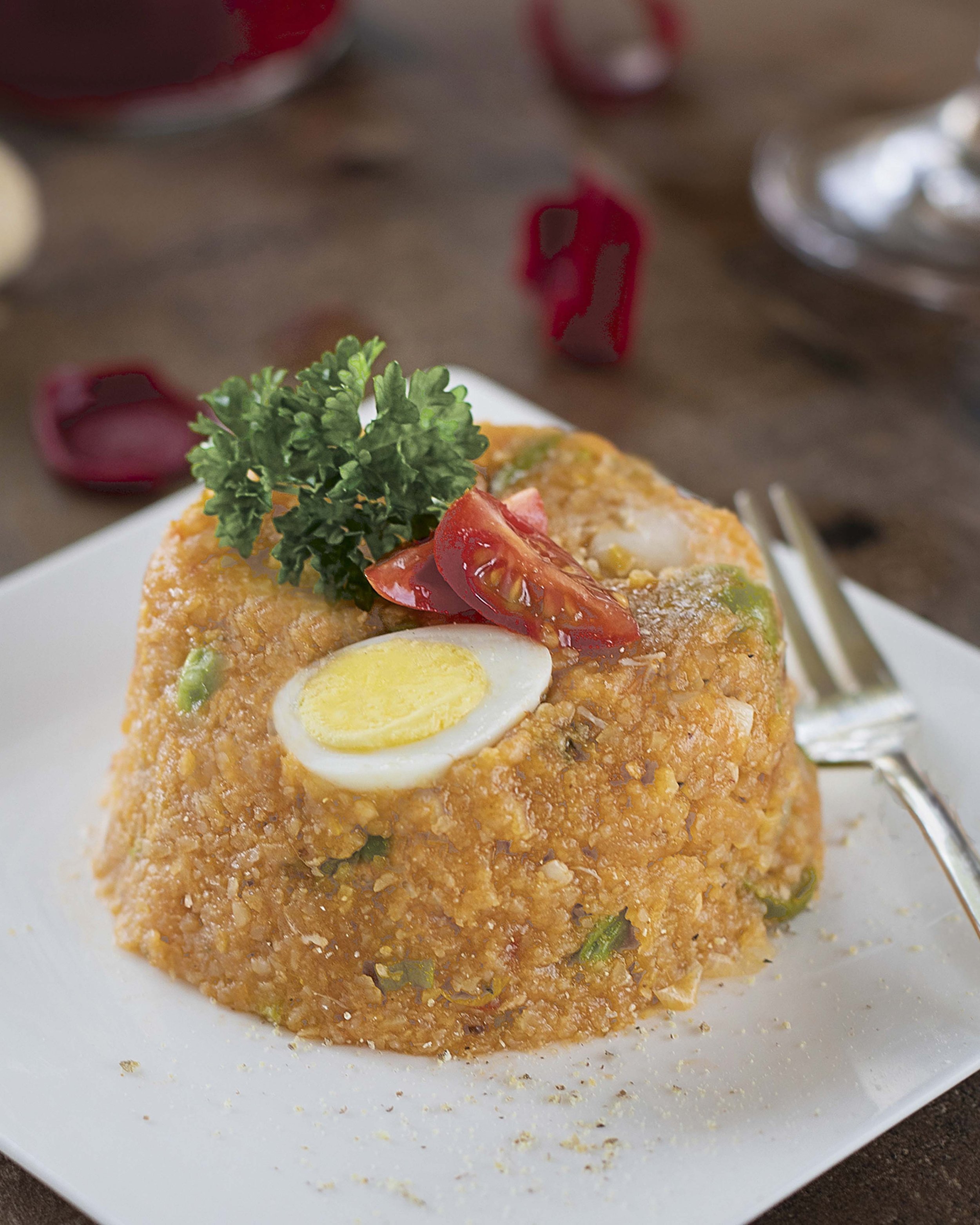 Close-up of a serving of rice topped with a halved boiled egg, cherry tomato slices, a sprig of parsley, and small green vegetable pieces on a white plate, with a fork beside it.