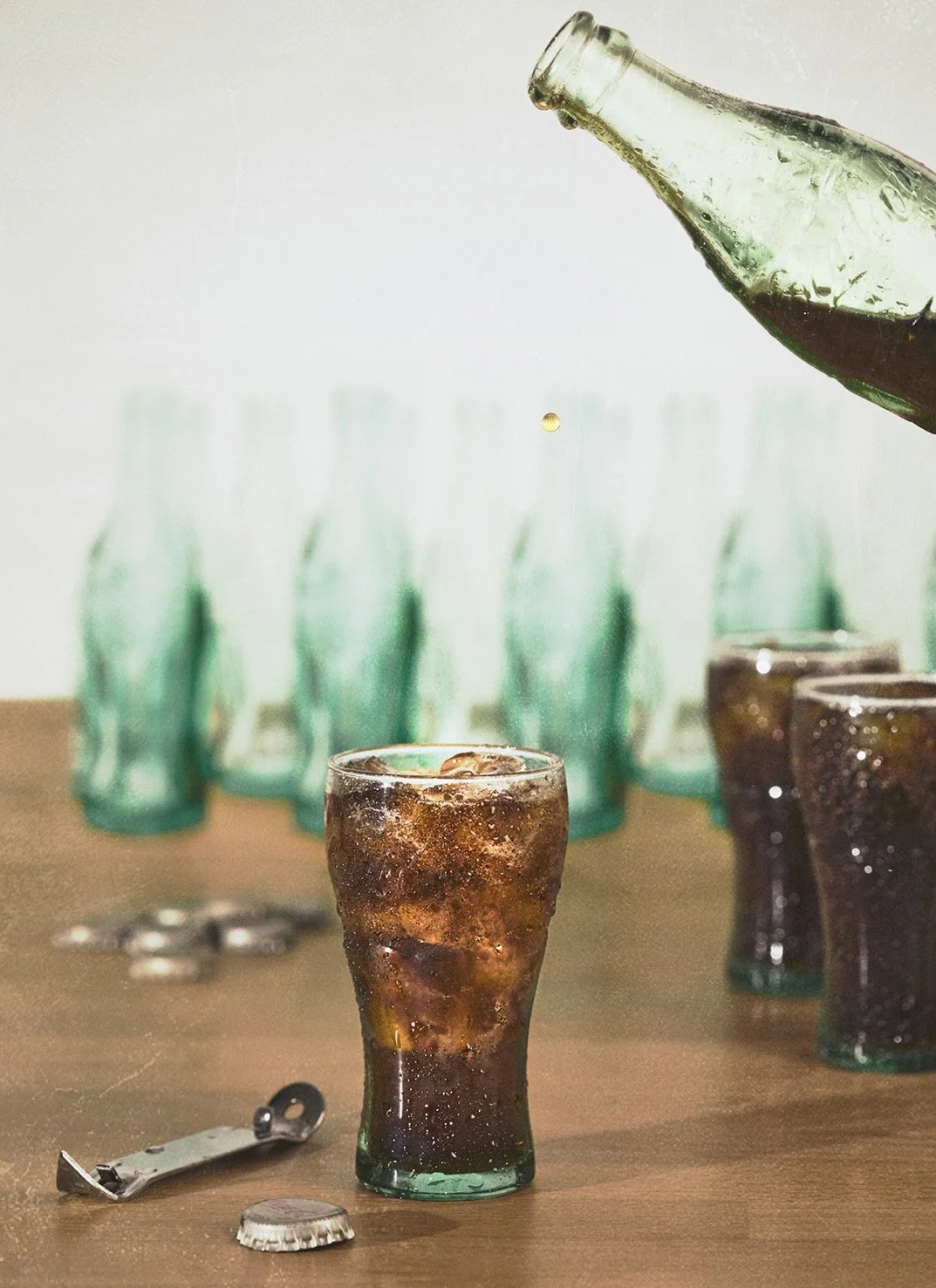 A glass of soda with ice on a table, with multiple green glass bottles in the background and an upside-down bottle pouring soda into the glass.